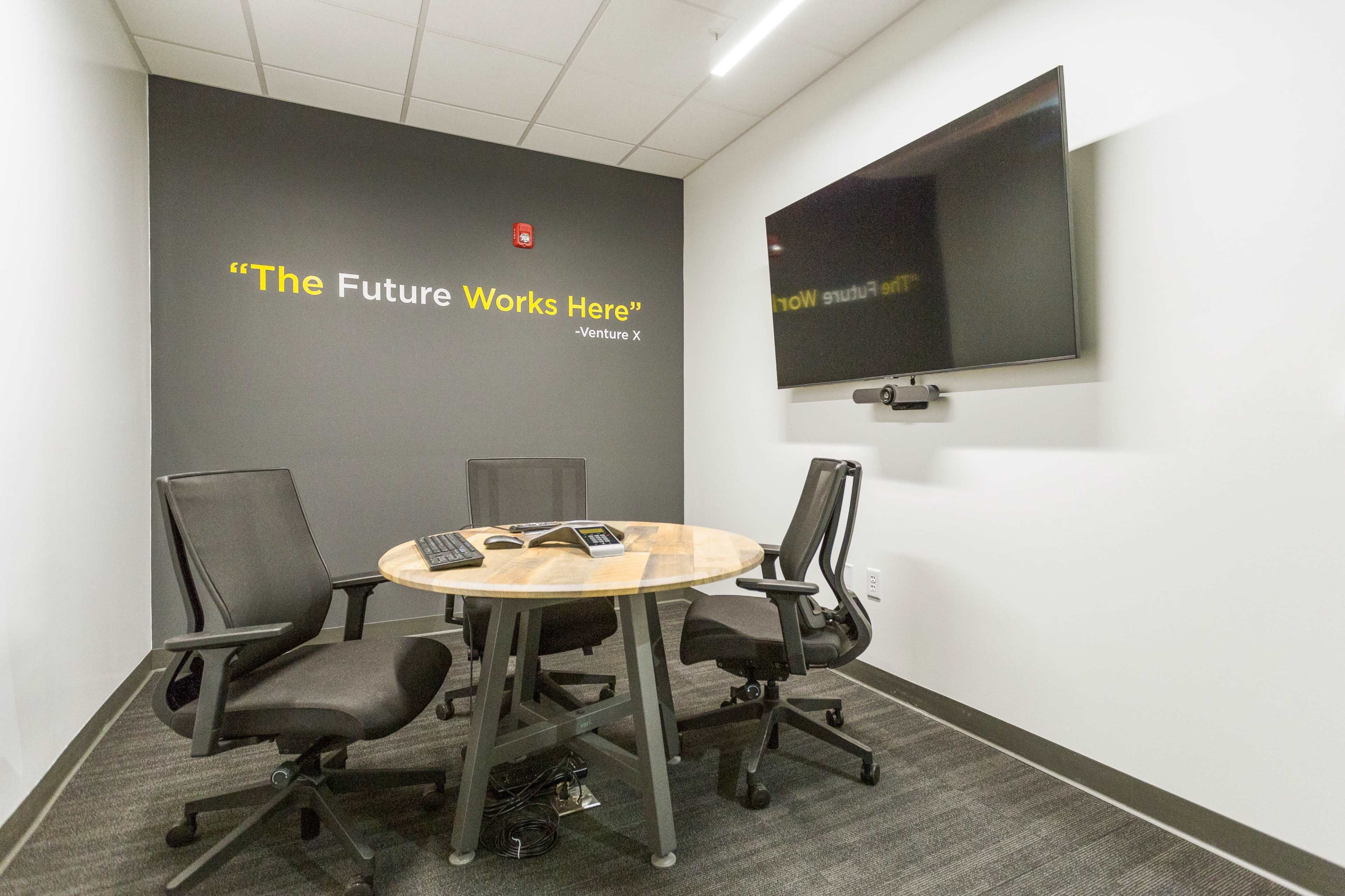 A small conference room features a round wooden table with three black chairs, a wall-mounted television, and a quote displayed on the wall.