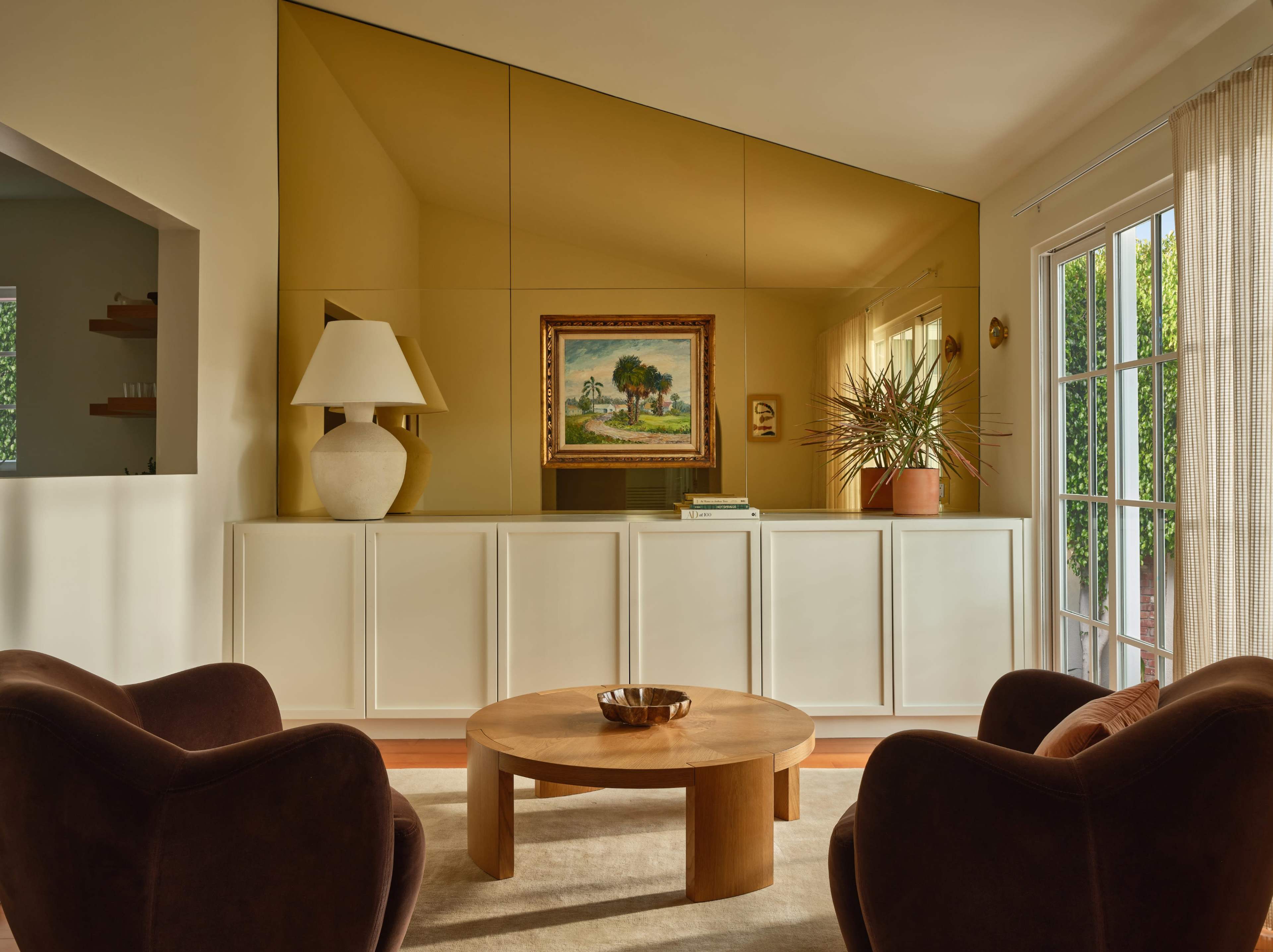 A modern living room features two brown armchairs facing a circular wooden coffee table, with a large golden mirror wall and a framed painting displaying a landscape.