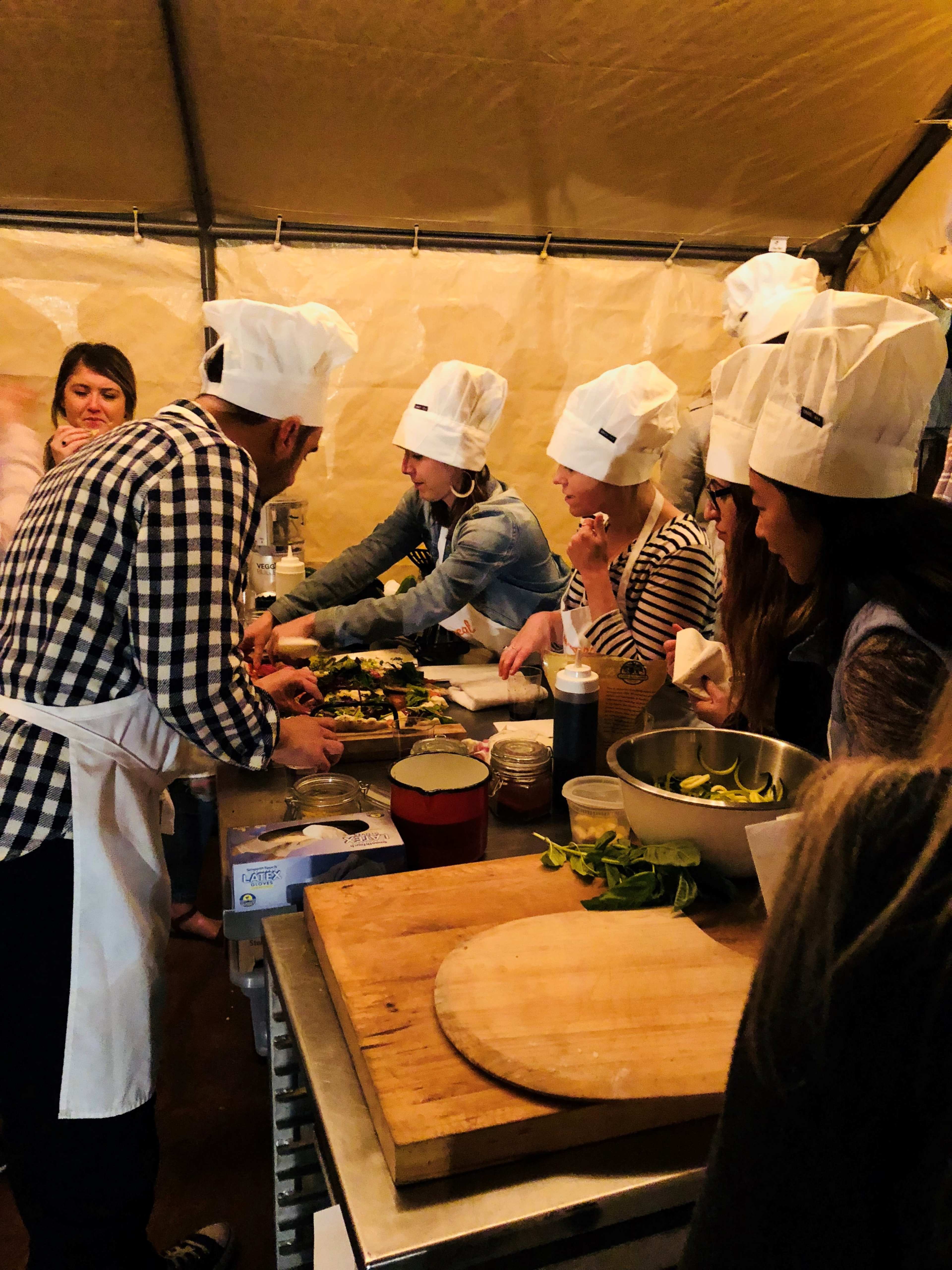 A group of people wearing chef hats prepares food in a tented kitchen.