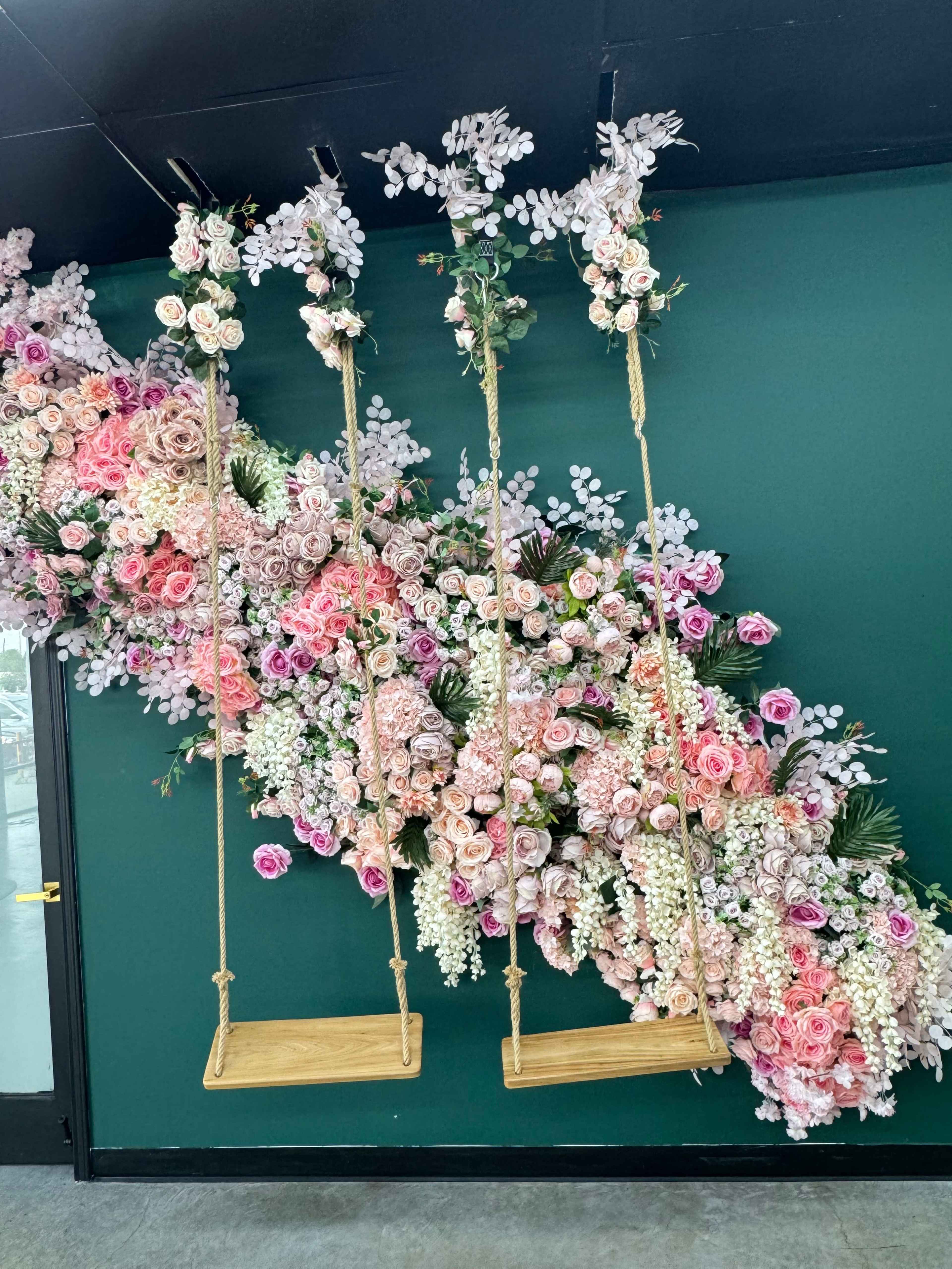 The image shows three wooden swings hanging from a floral installation featuring various pink and white flowers against a green wall.