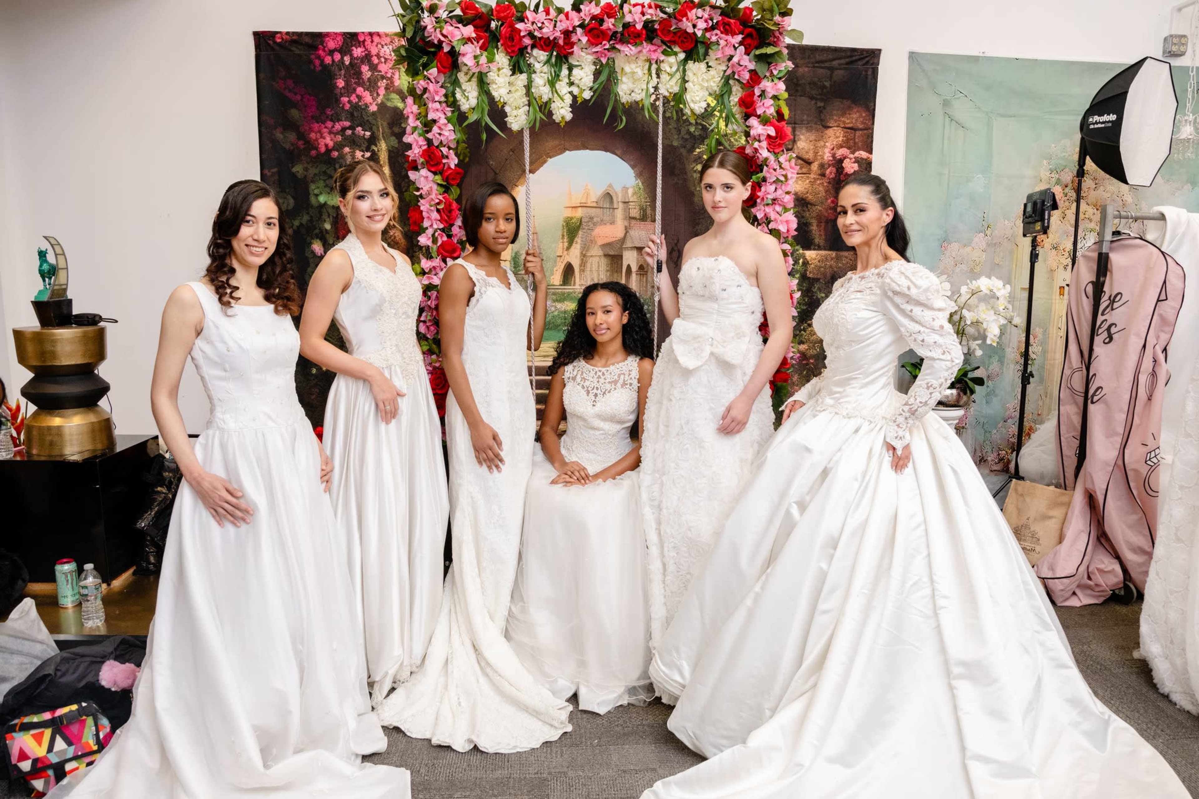A group of six models wearing white wedding gowns poses together in front of a floral backdrop.