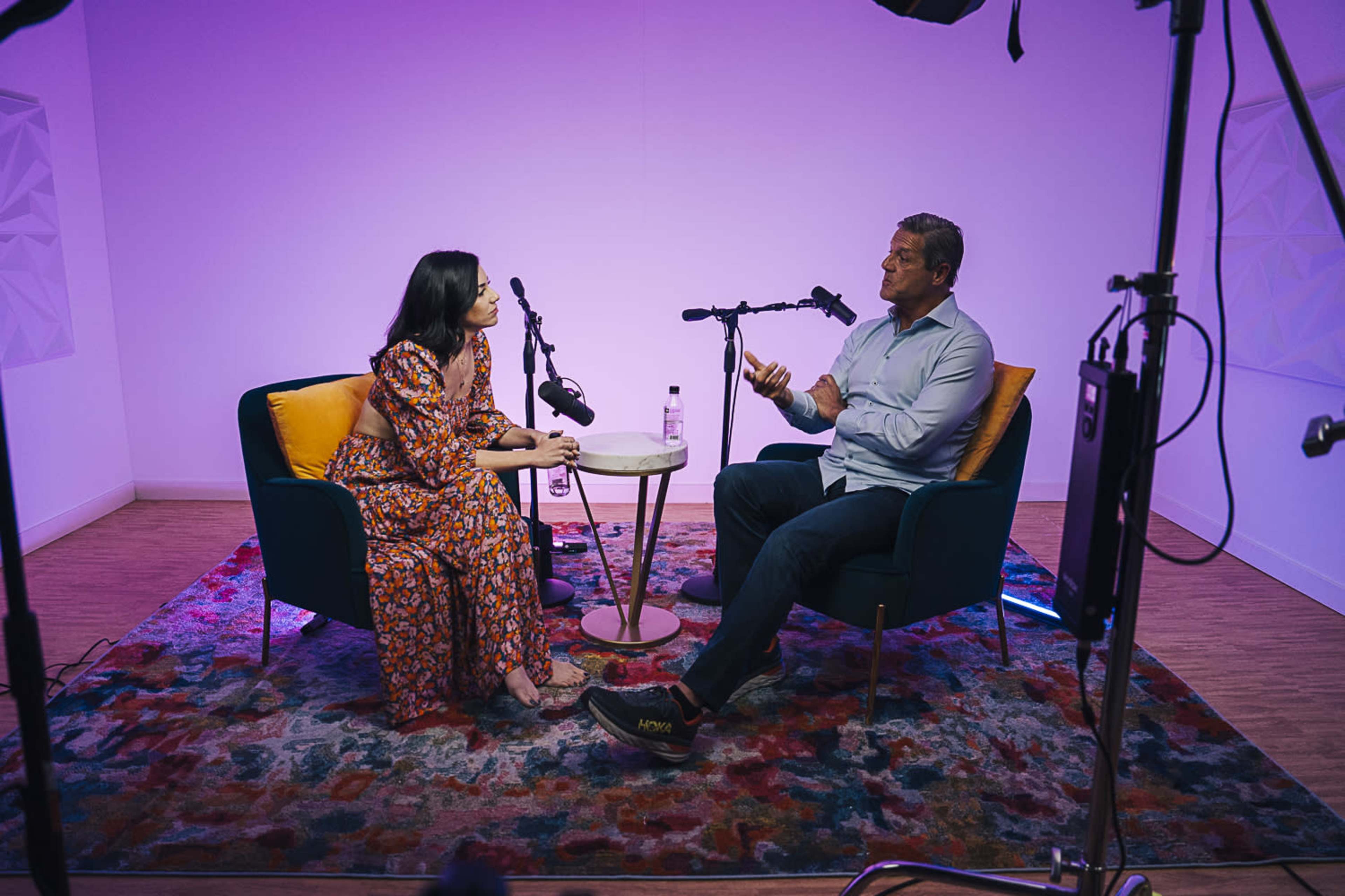 A man and a woman are seated in armchairs across from each other in a brightly lit studio, engaged in conversation with microphones in front of them.