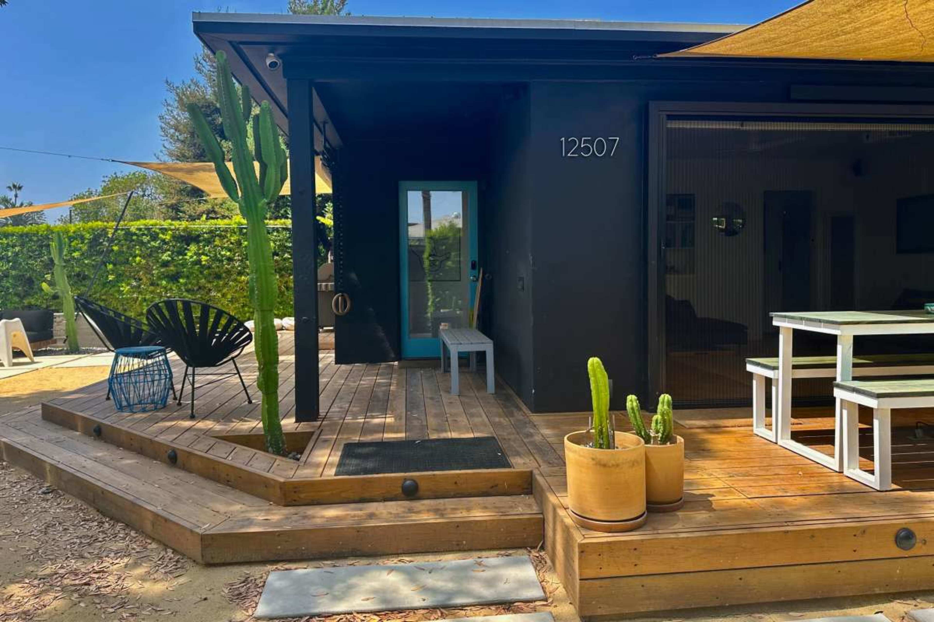 The image shows a modern single-story home with a wooden deck, a small blue bench, potted cacti, and a dining table situated outside.