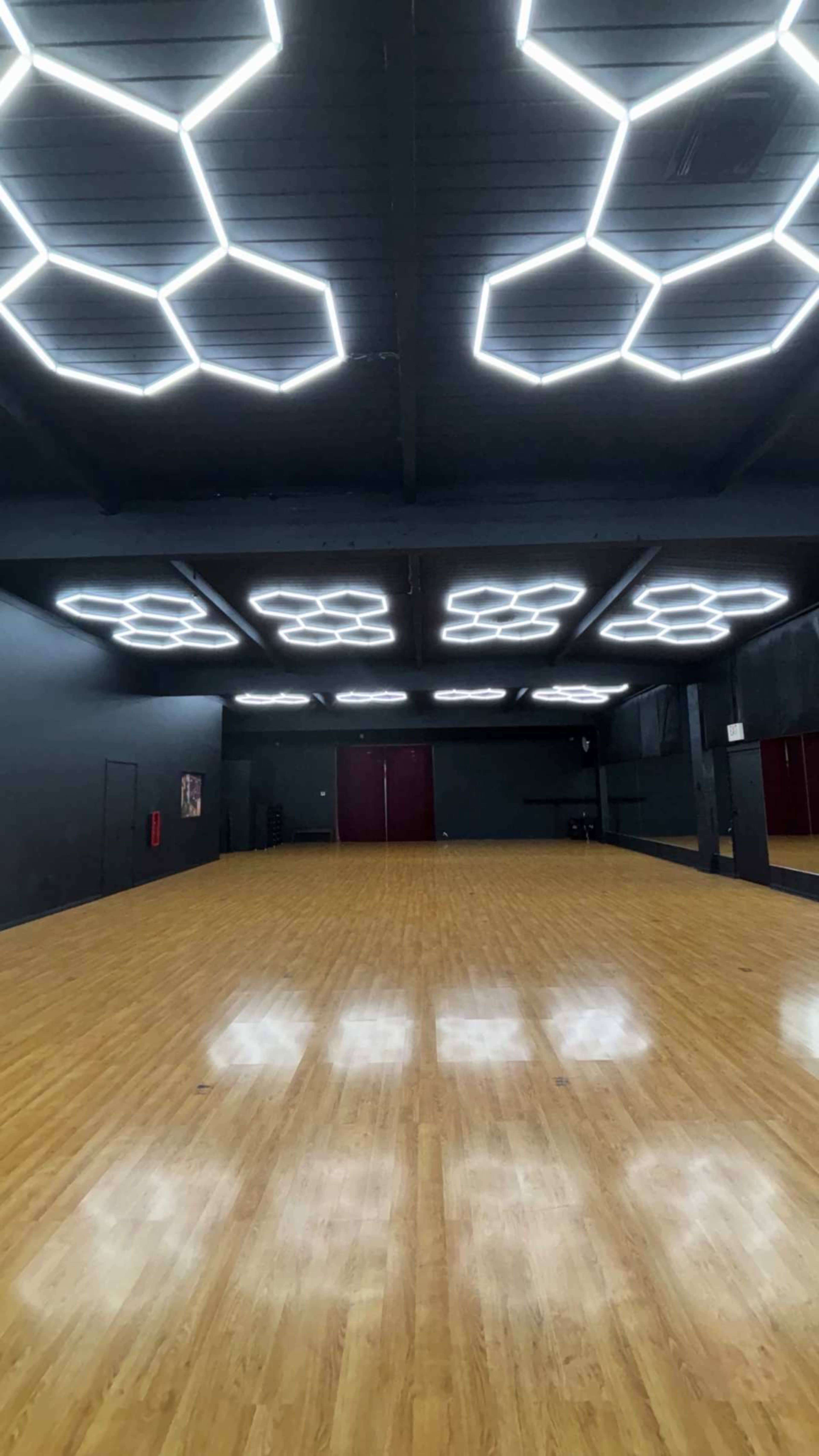 The image shows a spacious dance studio with a polished wooden floor and a ceiling featuring geometric hexagonal LED lights.