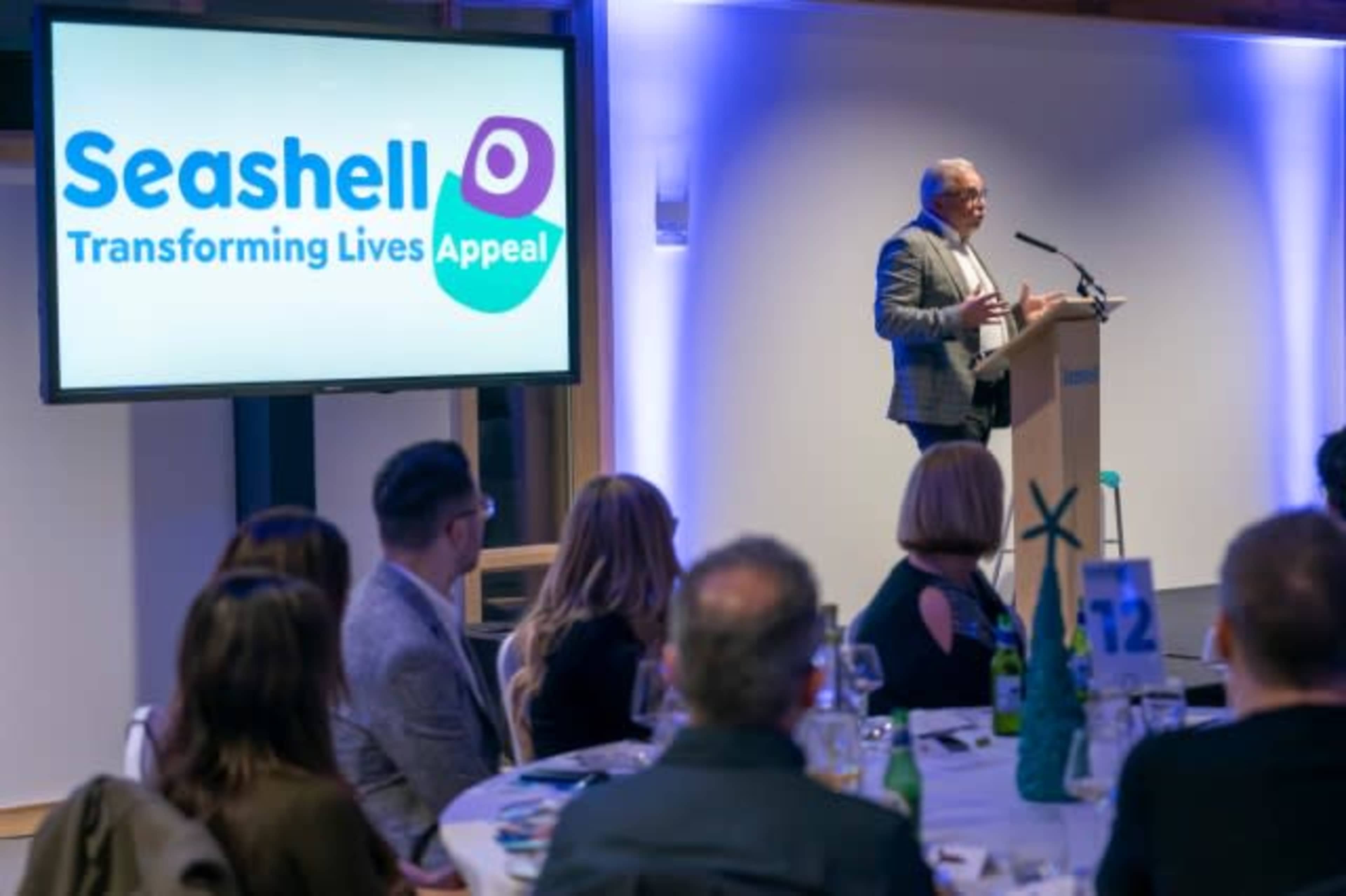 A speaker presents at a podium in front of an audience during an event for the Seashell Trust's Transforming Lives Appeal, with a large screen displaying the event's logo.