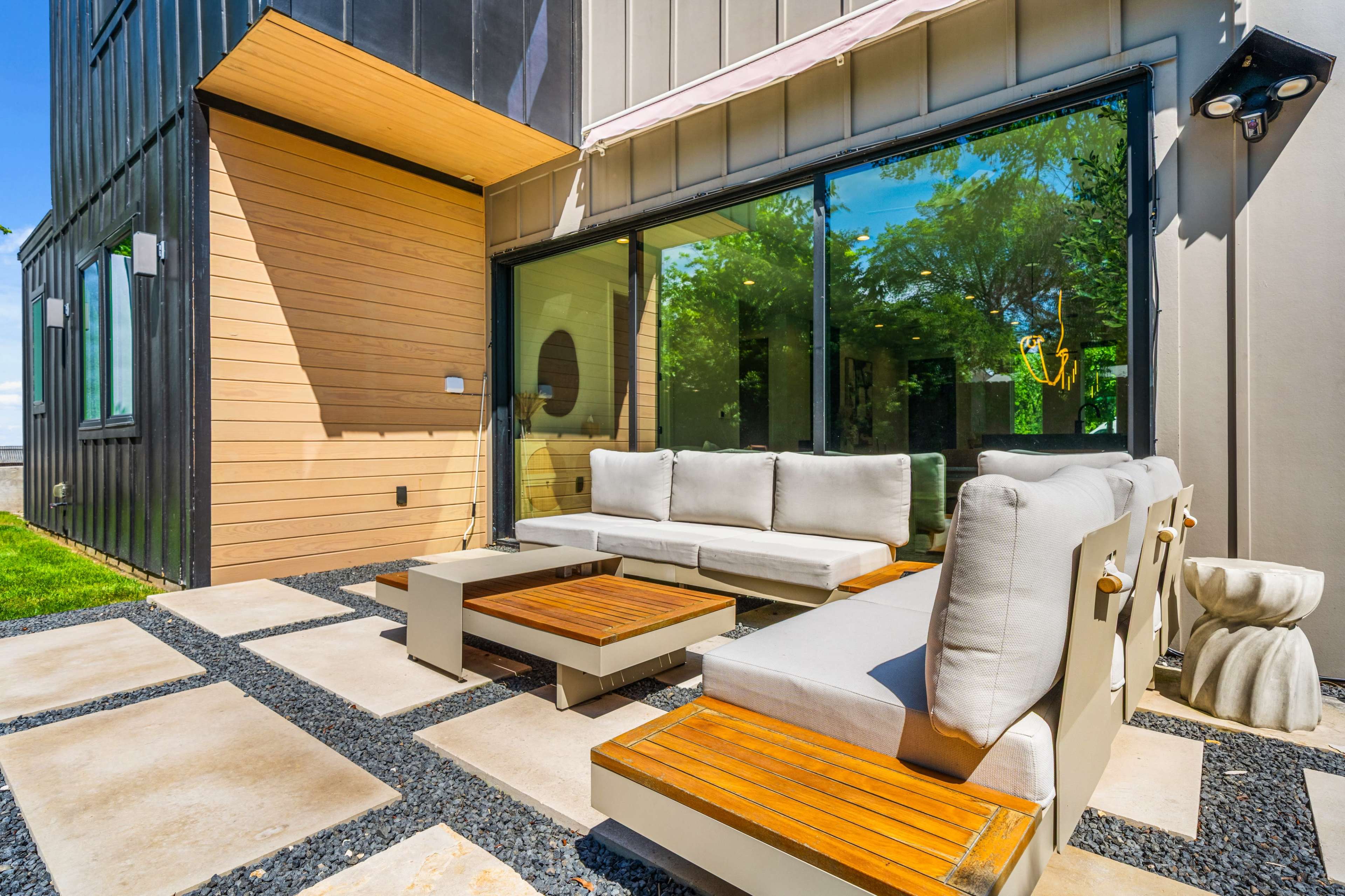 The image shows a modern outdoor patio with a seating area featuring a gray sectional sofa and wooden coffee tables, surrounded by stone pavers and gravel.