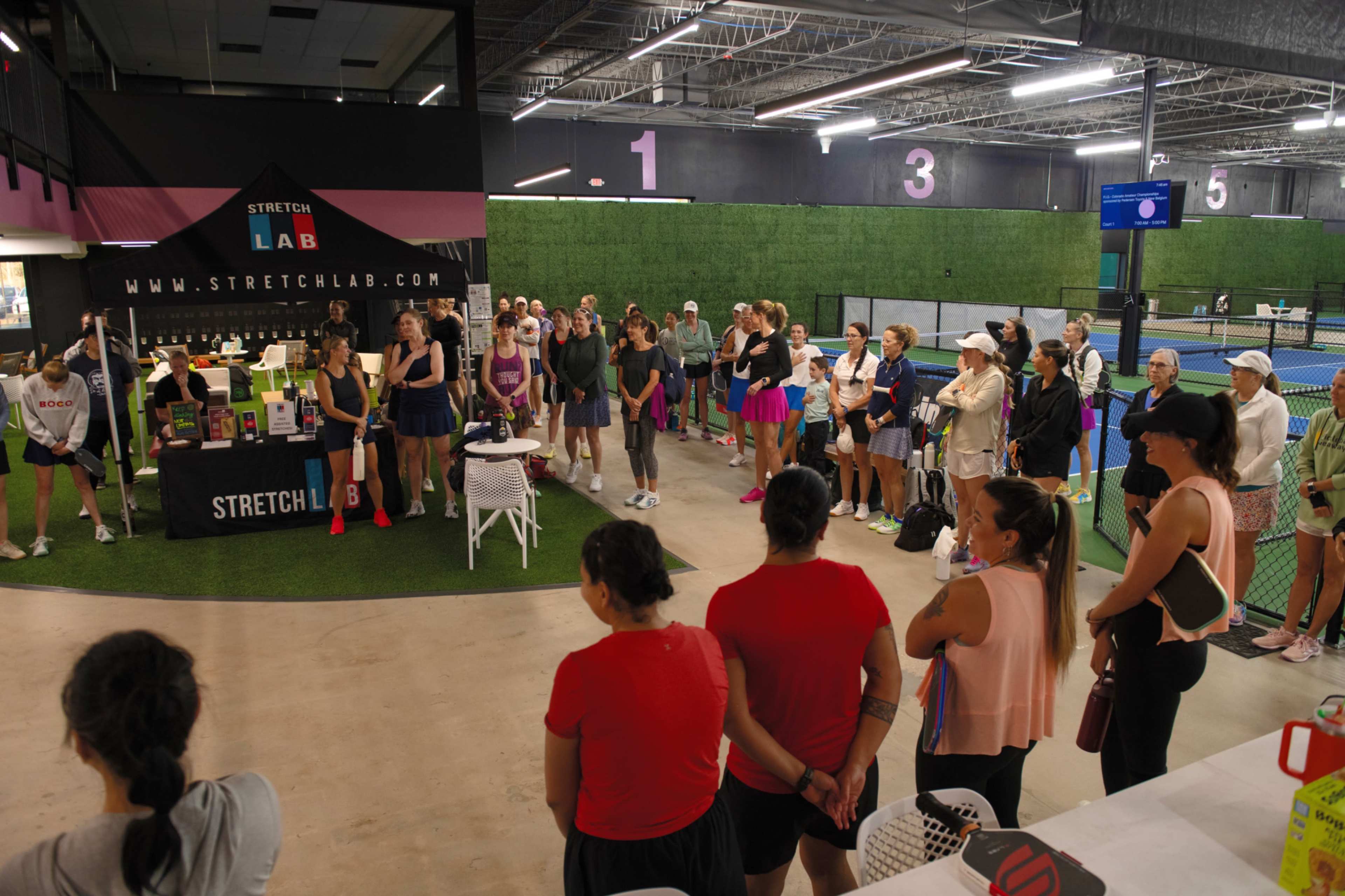 A group of people watches a presentation near a StretchLab booth at a sports facility.