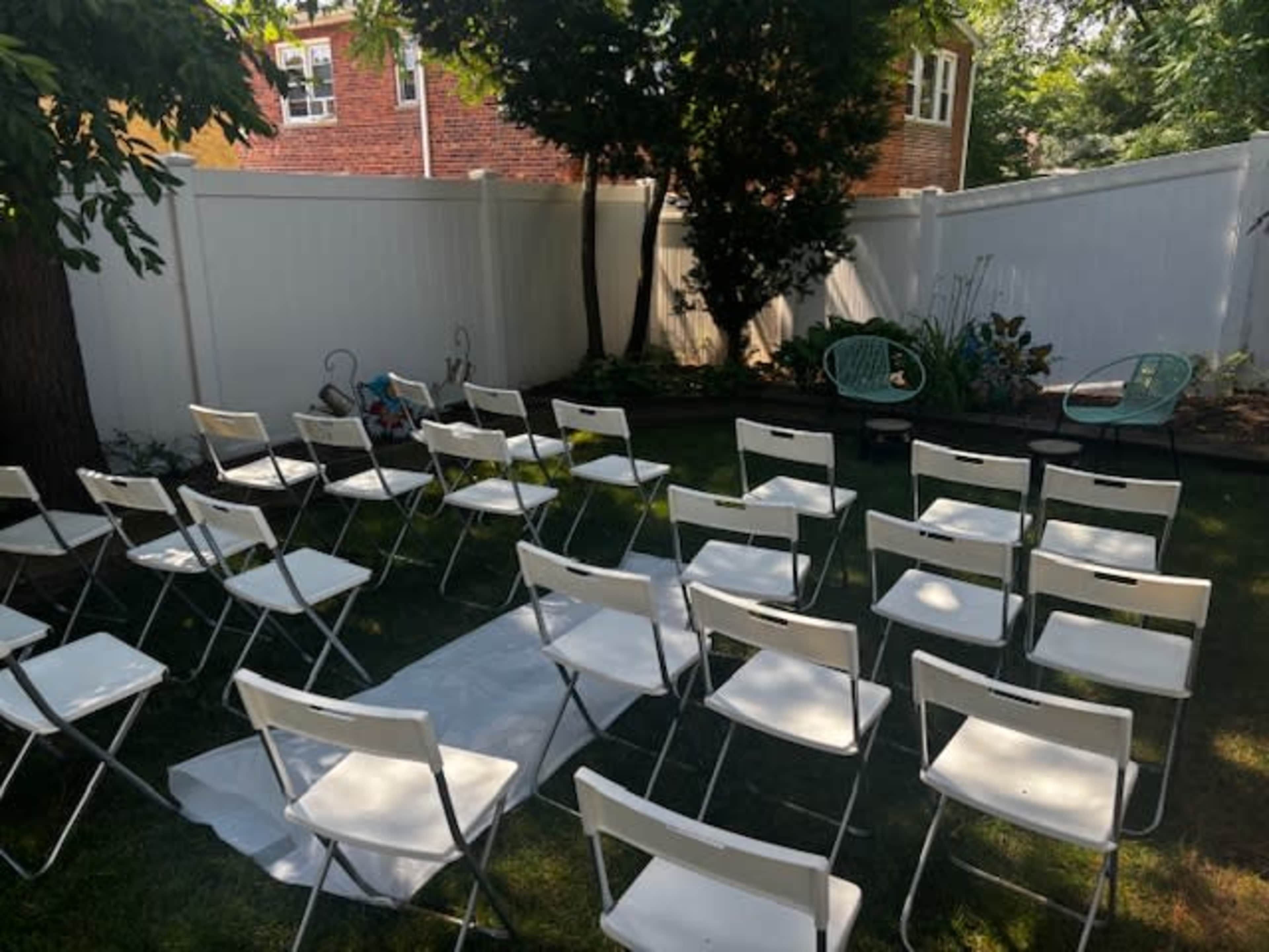 A backyard set up for an event, featuring white folding chairs arranged in rows facing a small path lined with a white runner, with greenery and decorative chairs in the background.