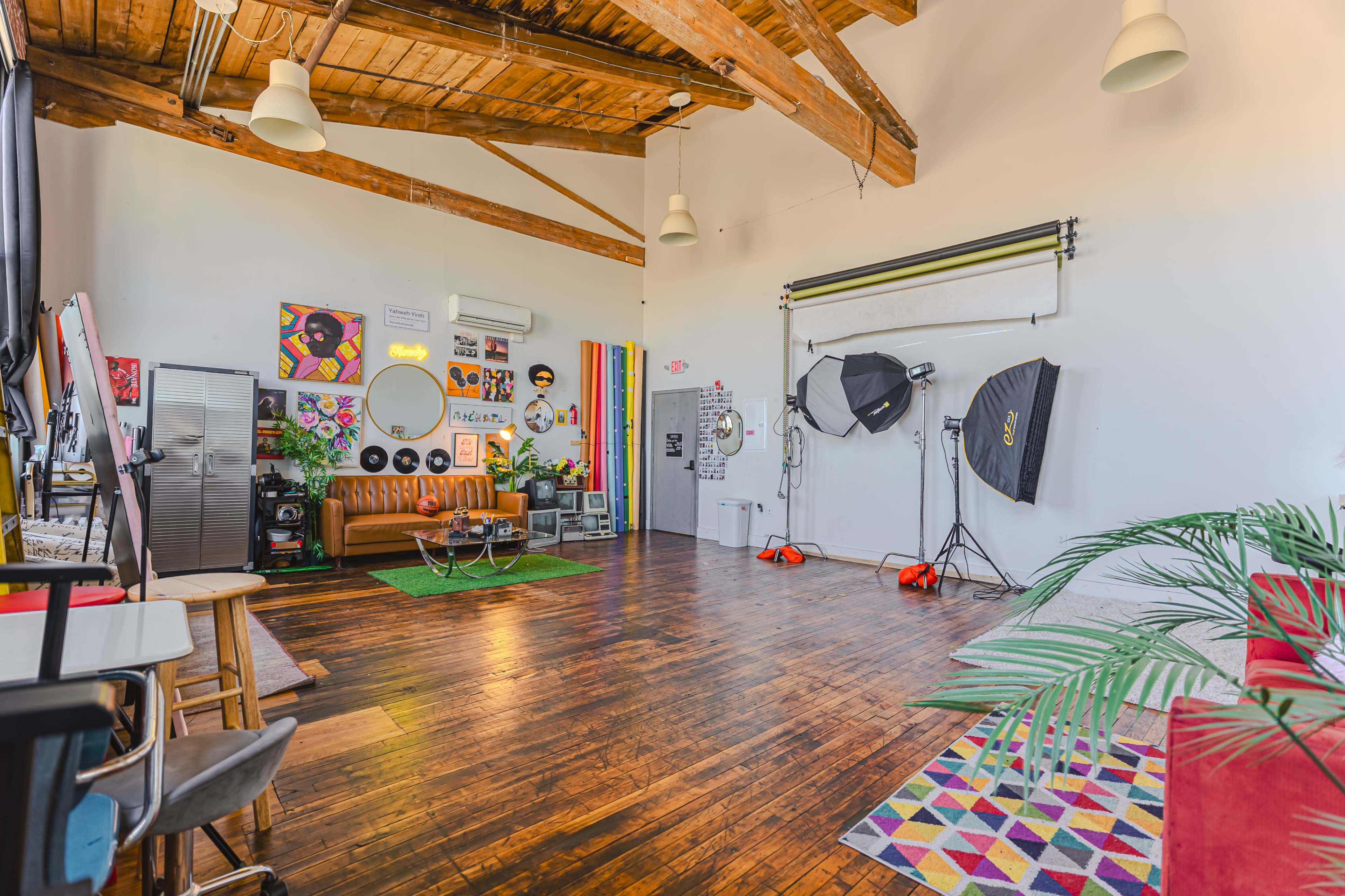The image shows a spacious, well-lit studio with wooden beams, a lounge area, photography equipment, and colorful decor.