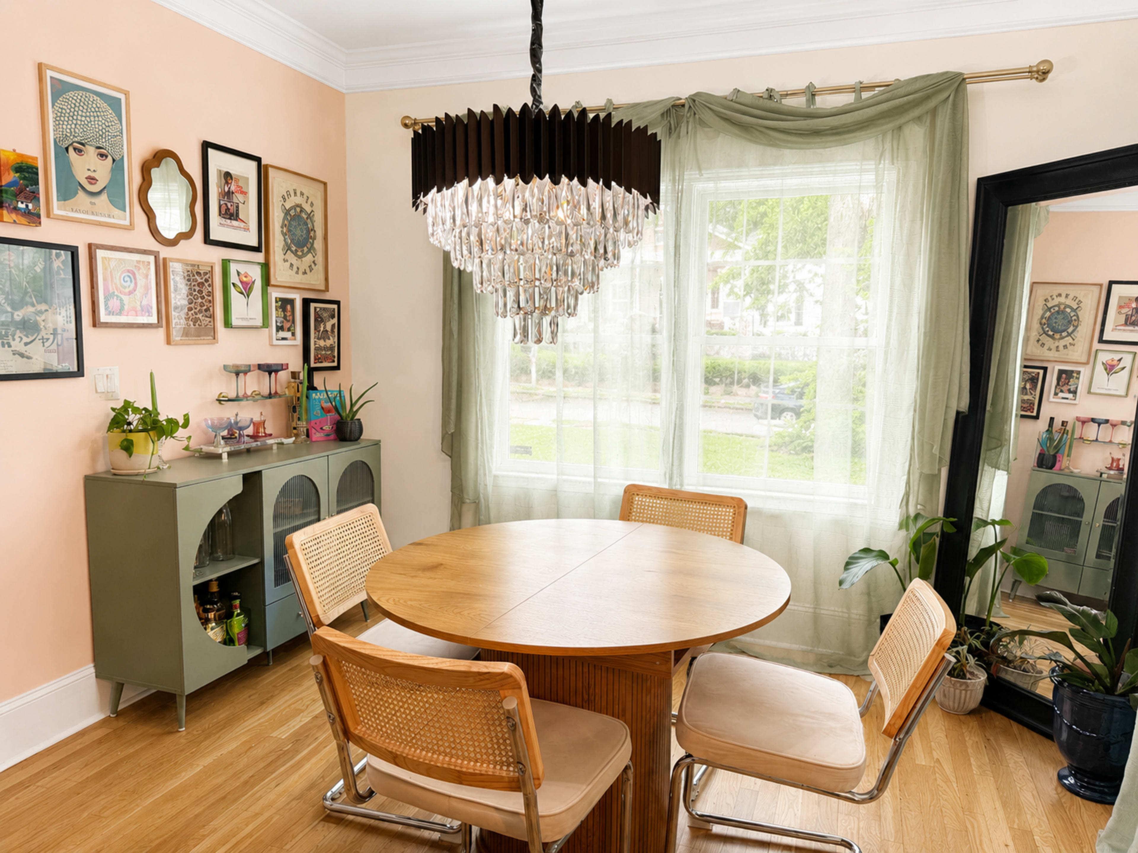 The image shows a dining area with a round wooden table surrounded by four chairs, elegant artwork on the walls, and a prominent chandelier hanging above the table.