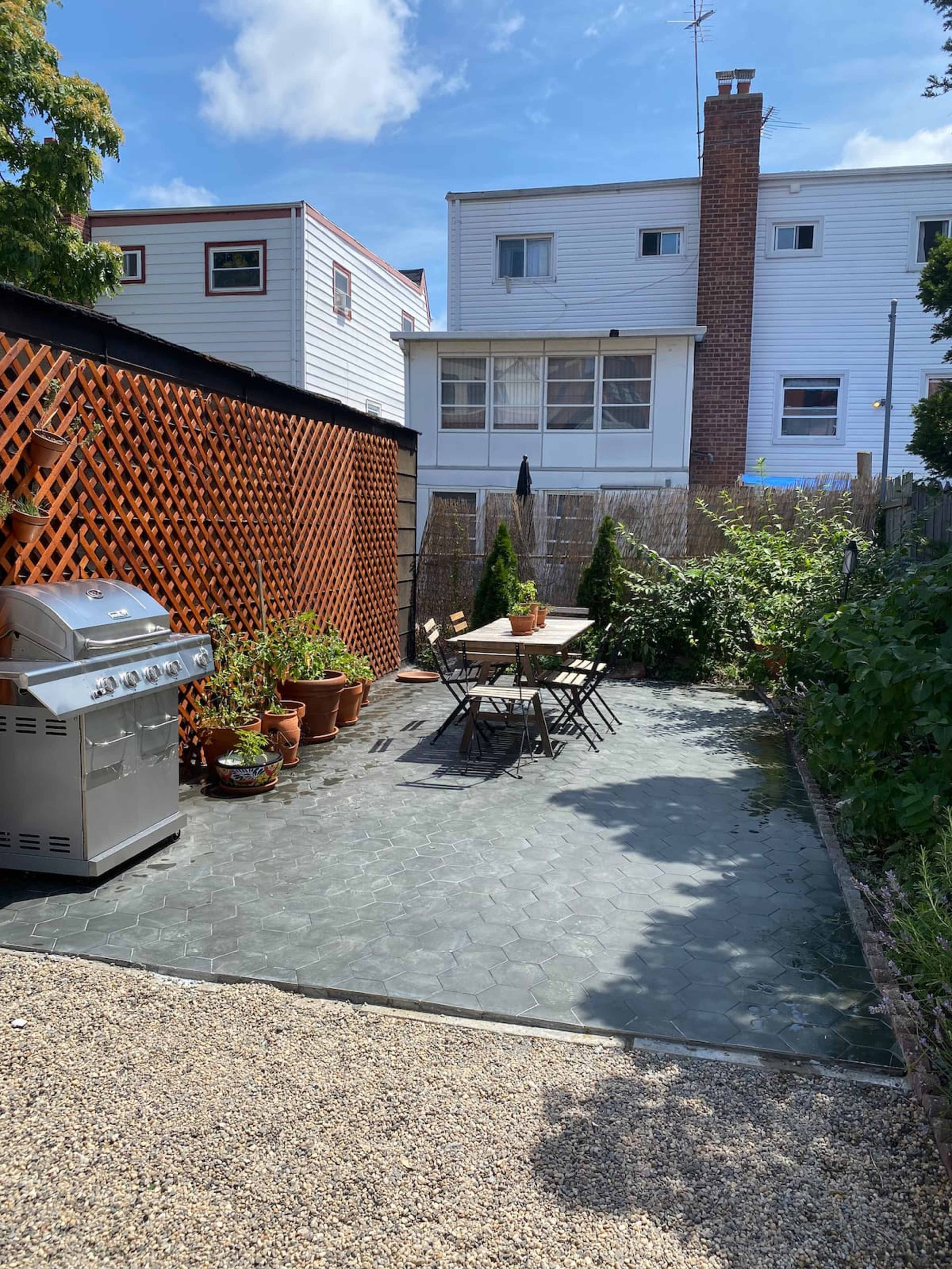 A small outdoor patio area with a table and chairs, a barbecue grill, potted plants, and surrounding wooden fencing.