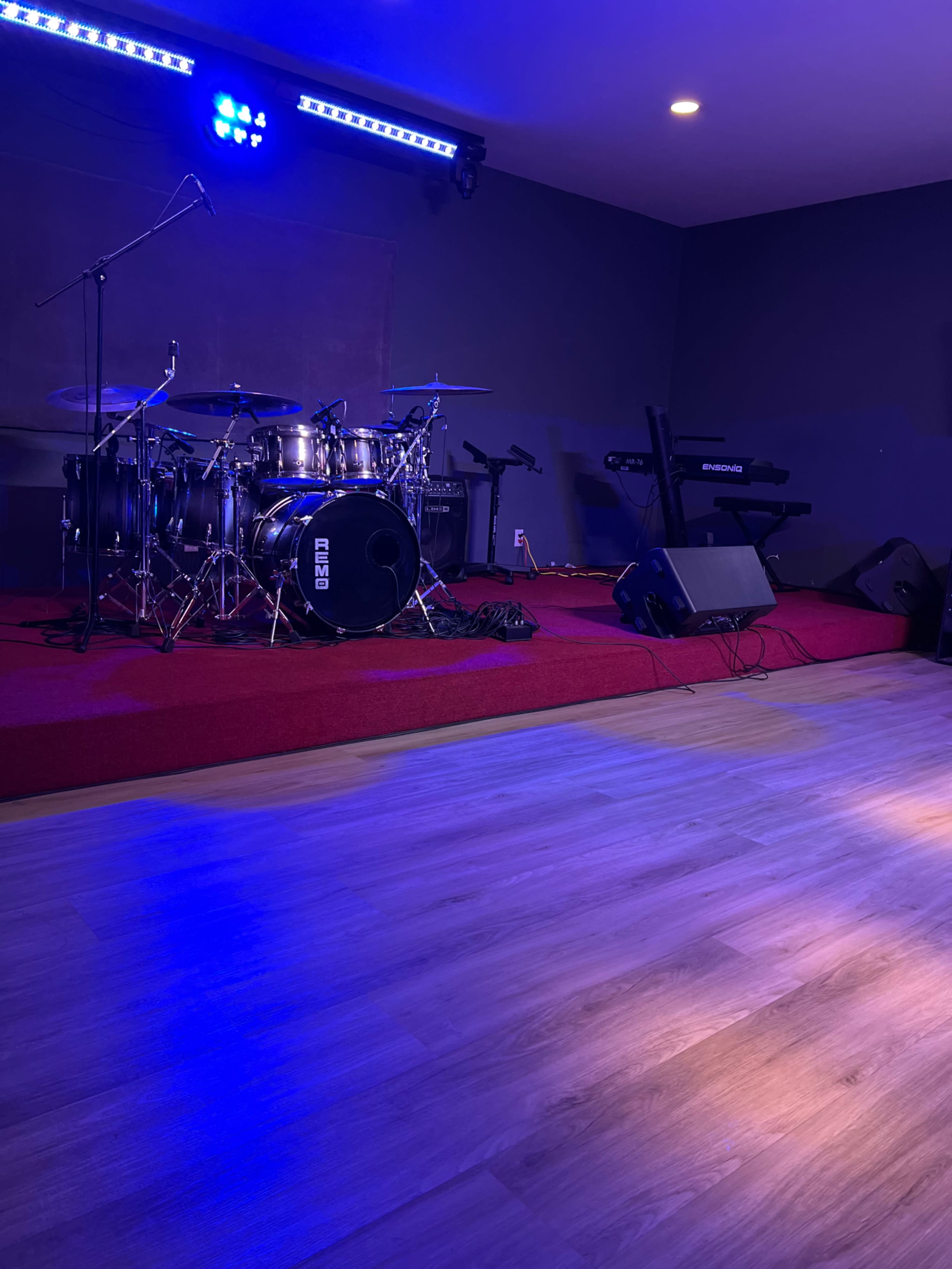 A drum set is positioned on a stage with red carpeting, accompanied by a keyboard and blue lighting illuminating the scene.
