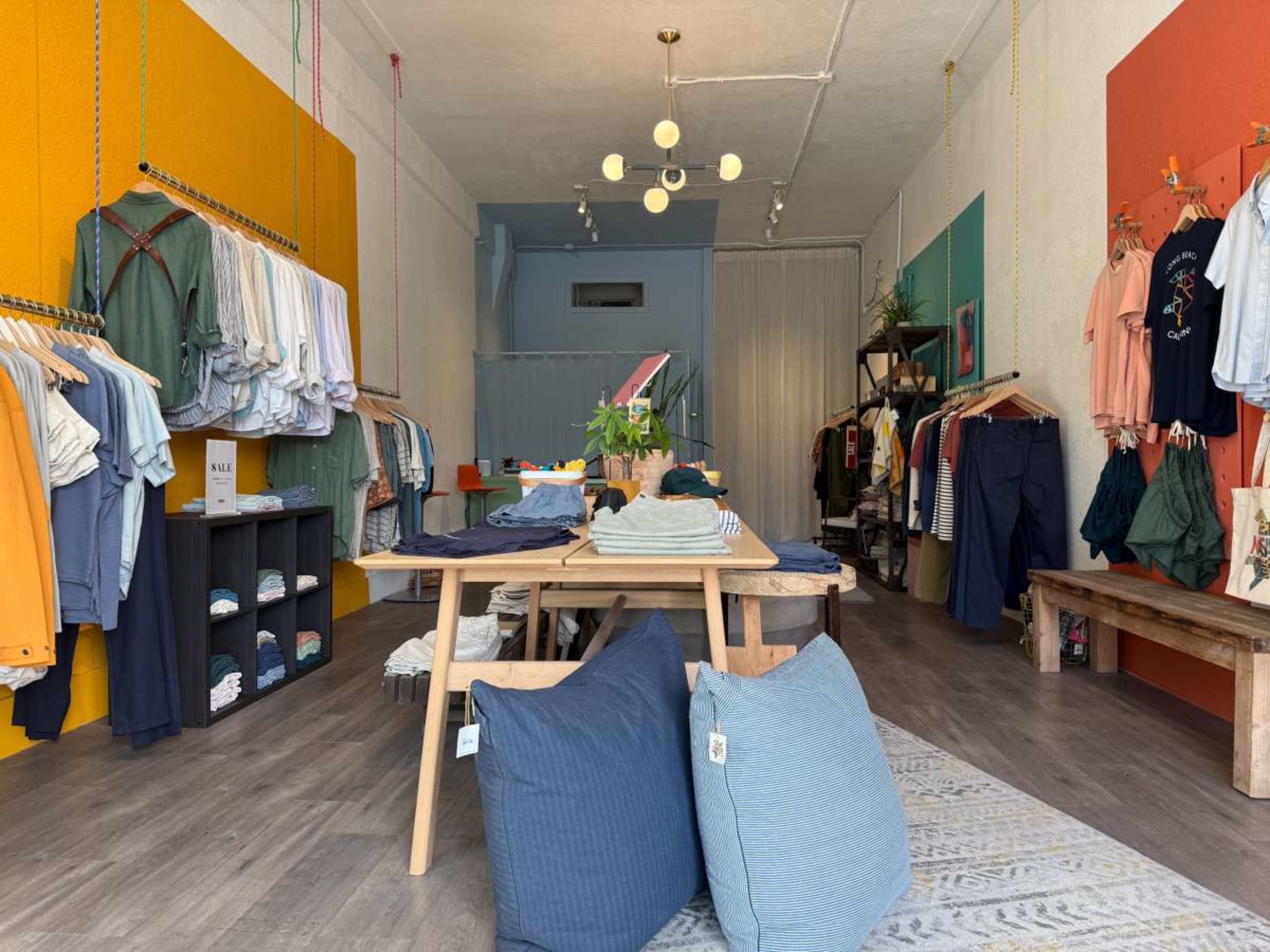 The image shows a clothing store with racks of apparel lining the walls, a display table in the center, and decorative pillows on the floor.