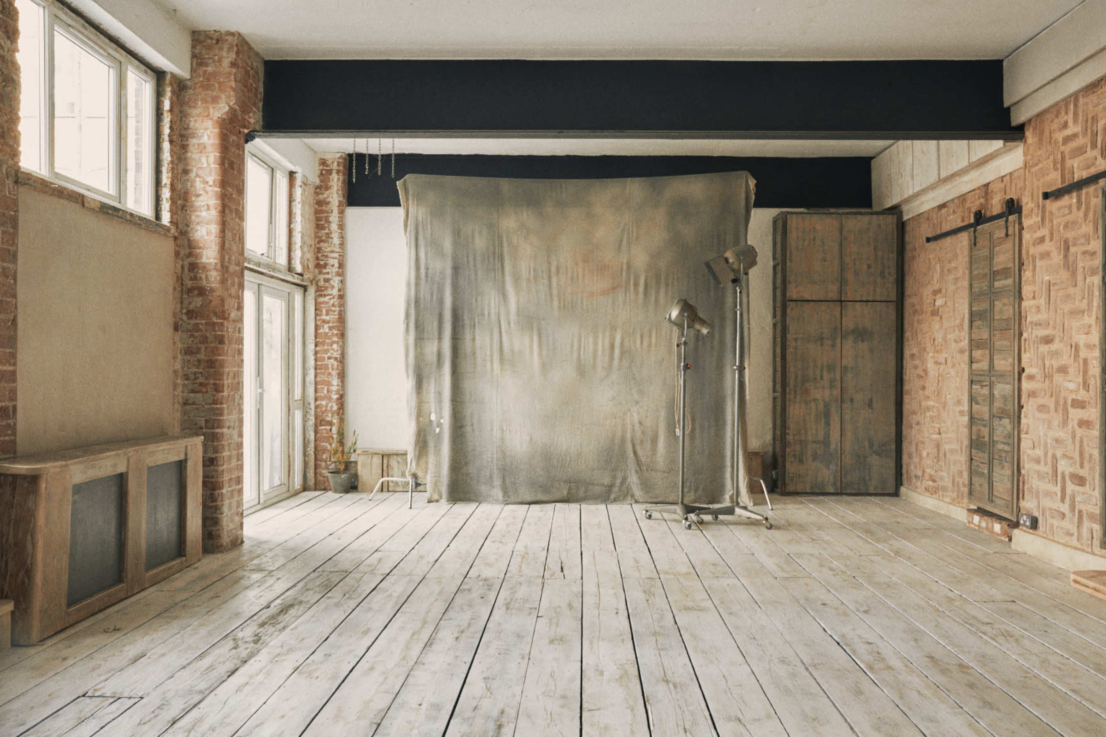 A spacious photographic studio with wooden floors, exposed brick walls, and a large fabric backdrop set up for shooting.