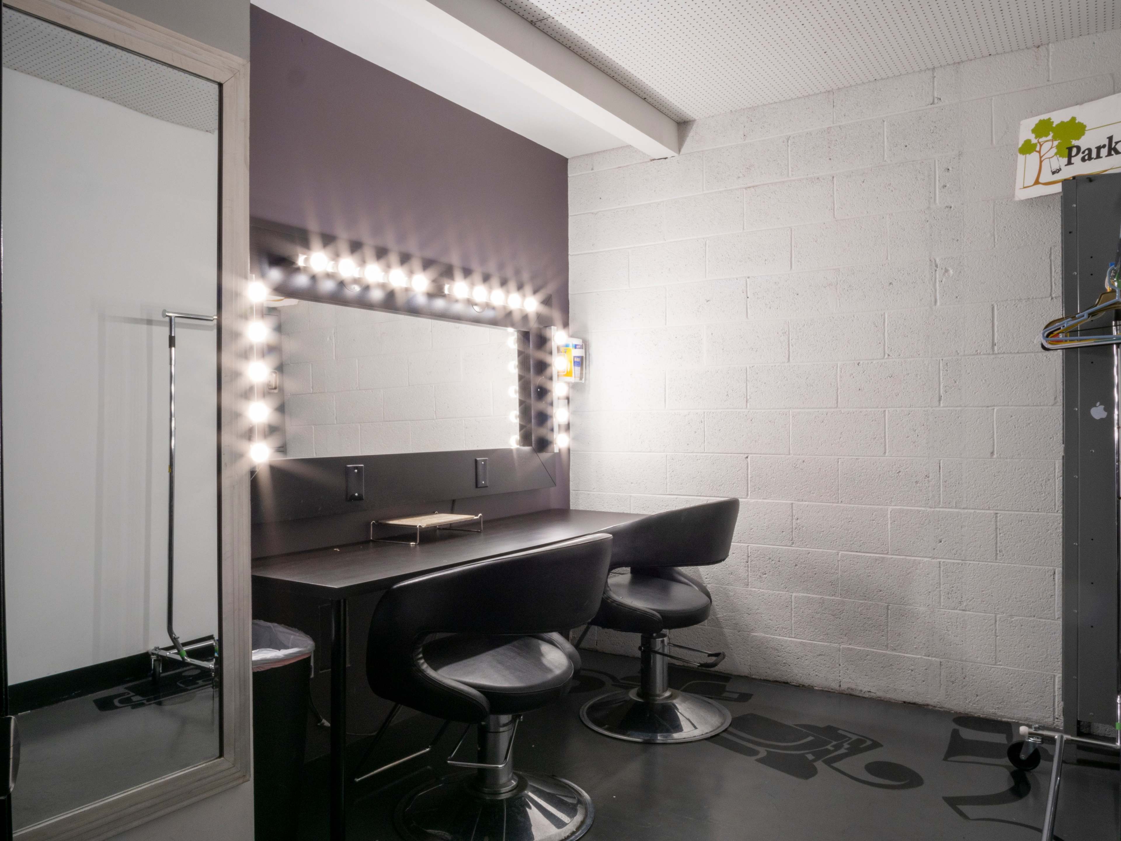 A minimalistic dressing area featuring a long table with two black chairs, a large illuminated mirror, and a wall with a hanging sign.