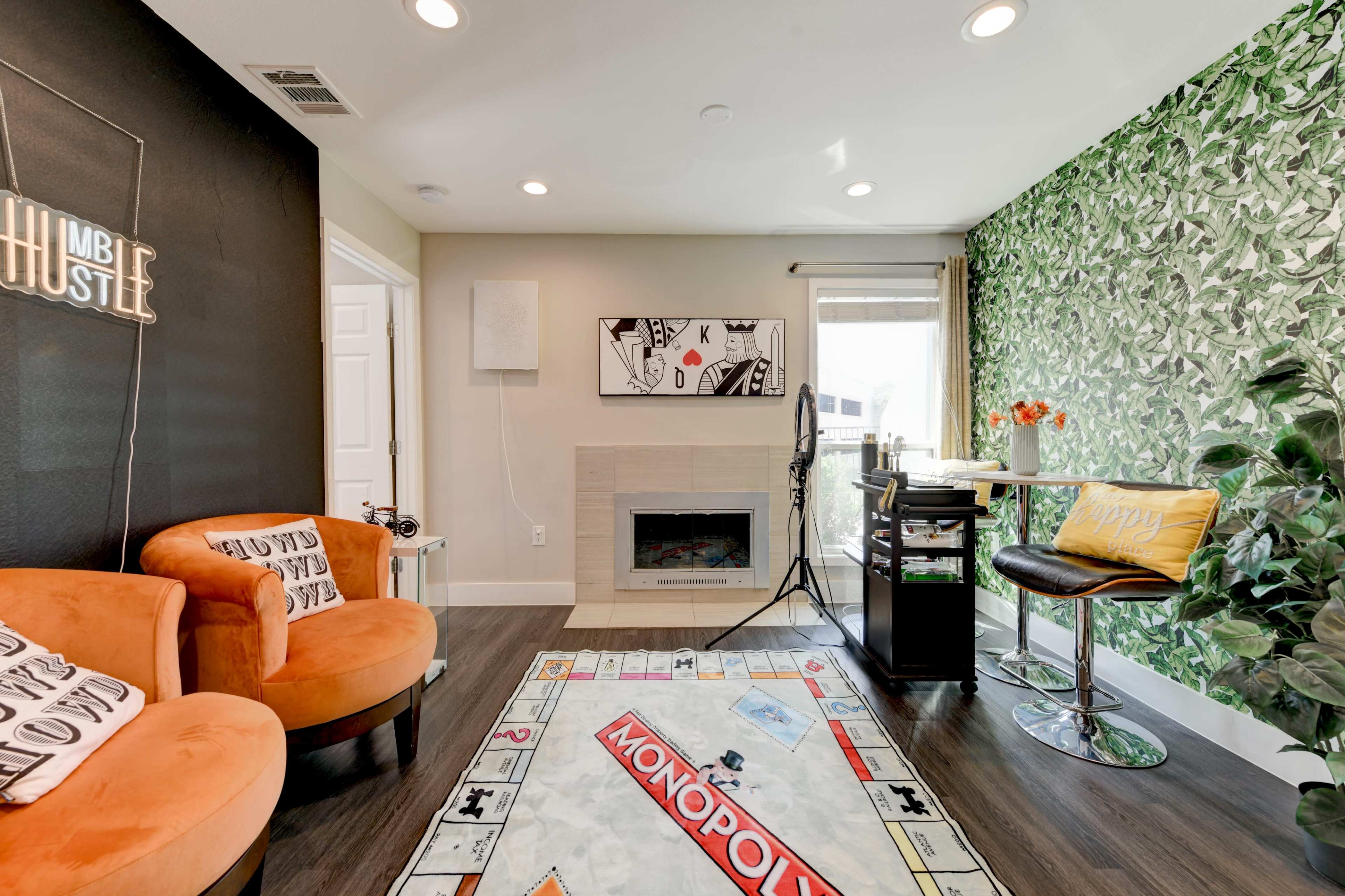 A cozy room featuring a green leafy accent wall, a fireplace, comfortable orange chairs, and a Monopoly-themed rug.