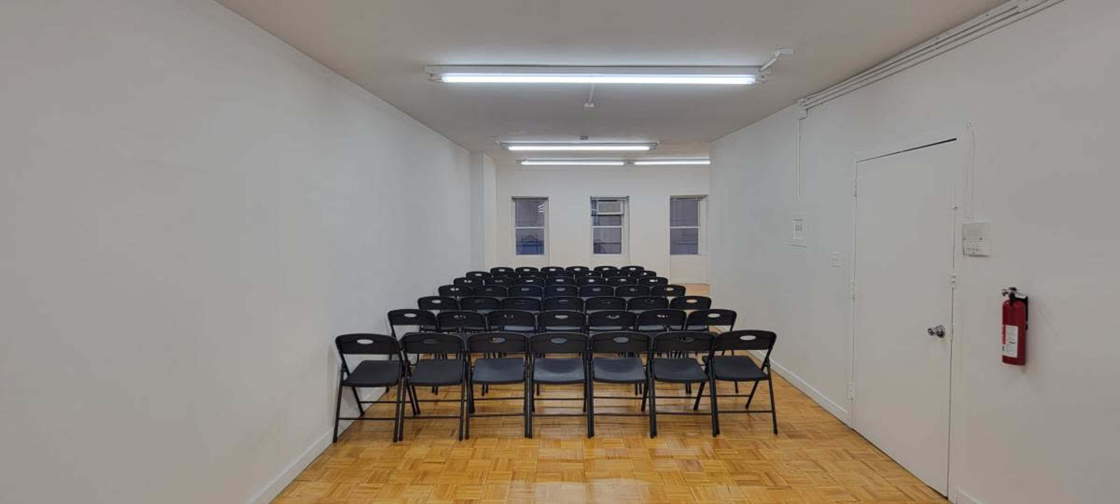 The image shows a row of black chairs arranged neatly in a spacious, empty room with white walls and wooden flooring.