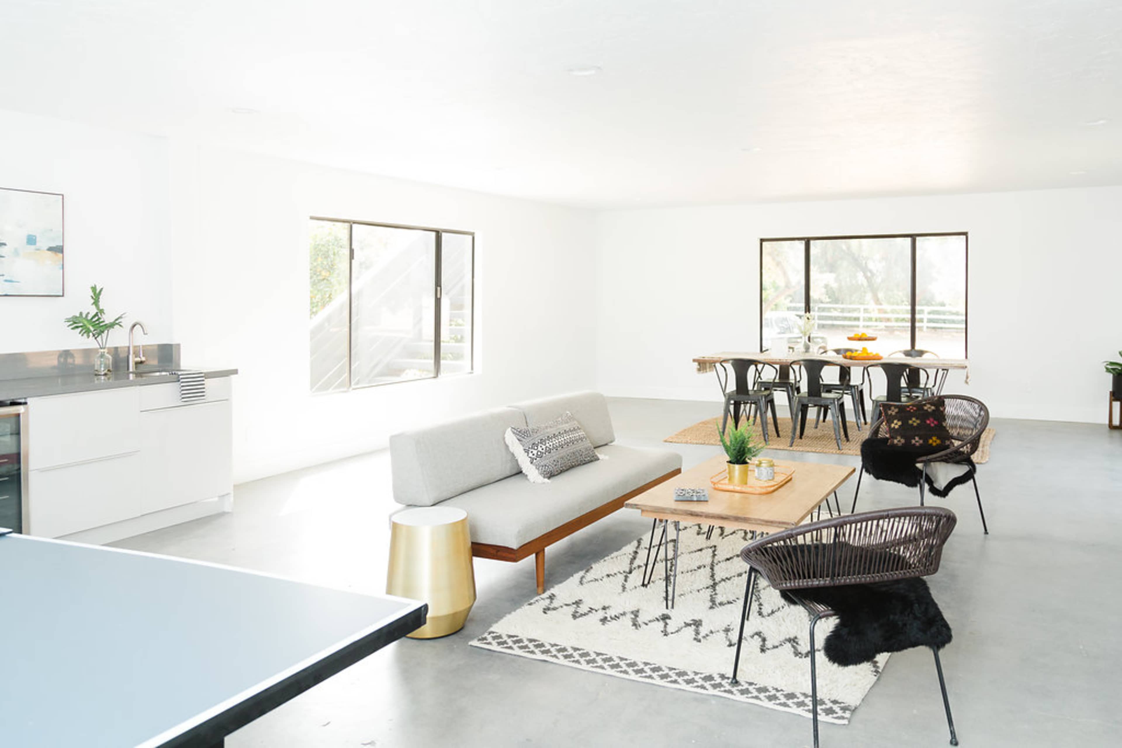The image shows a modern, open living space with a kitchen area, a seating arrangement featuring a gray sofa and wooden coffee table, and a dining table with chairs.