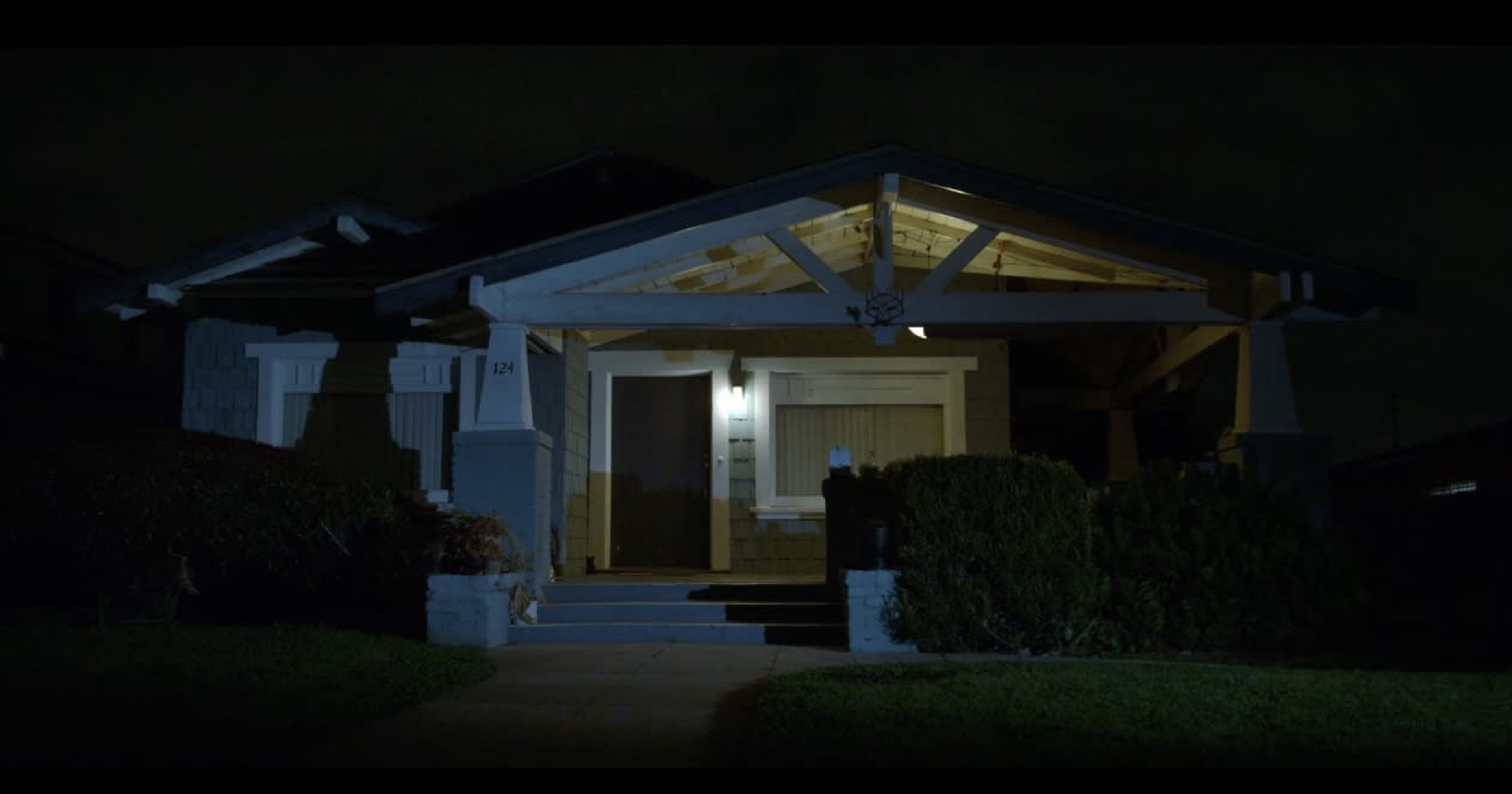 A darkened house with a front porch lit by a single light, surrounded by low hedges and a neatly manicured lawn.