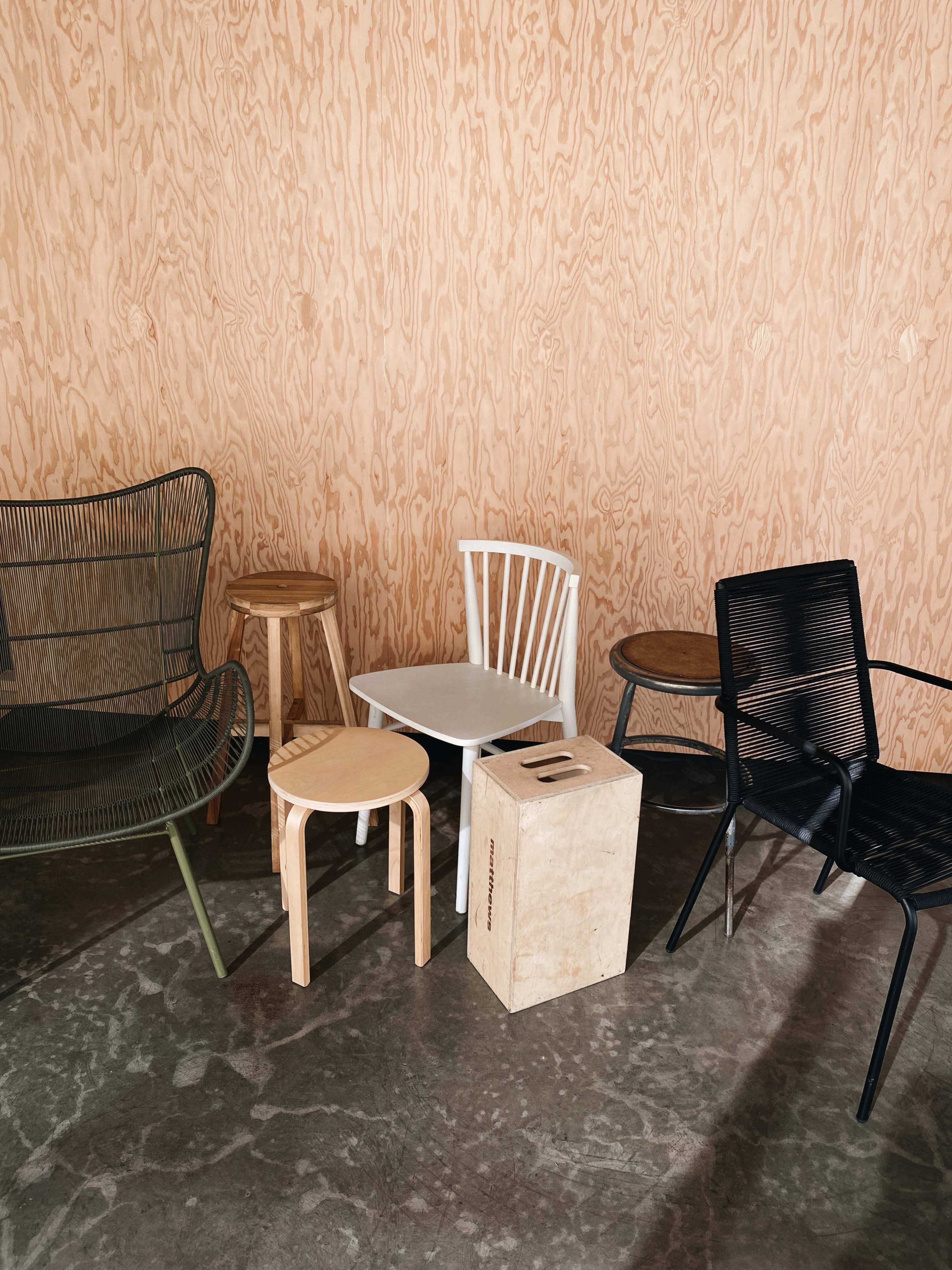 A variety of chairs and stools arranged on a concrete floor against a wooden wall.