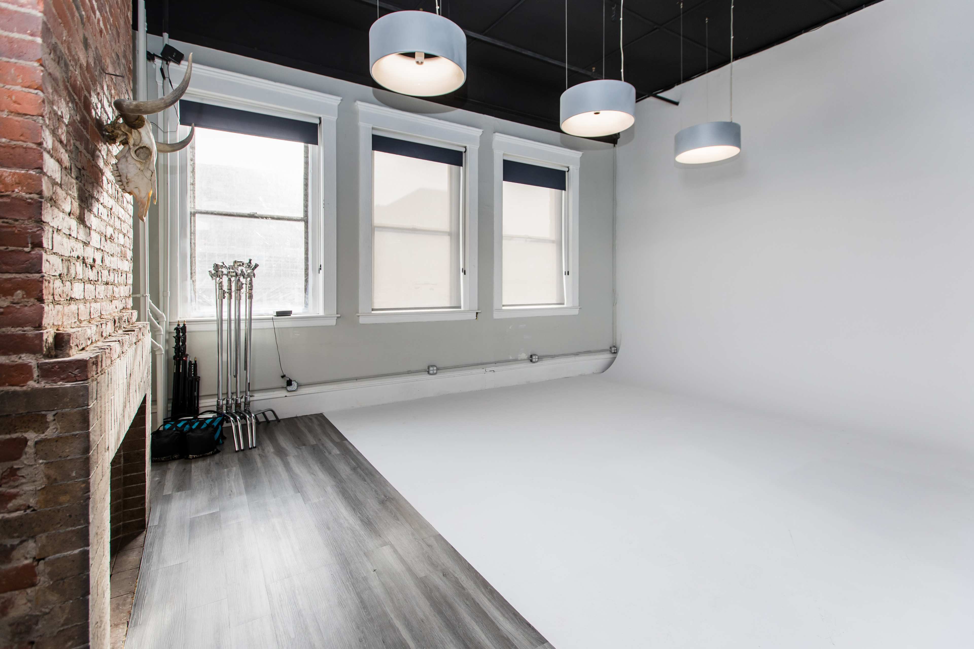 A spacious room with brick walls, large windows, and a white wall, featuring modern light fixtures and a wooden floor.