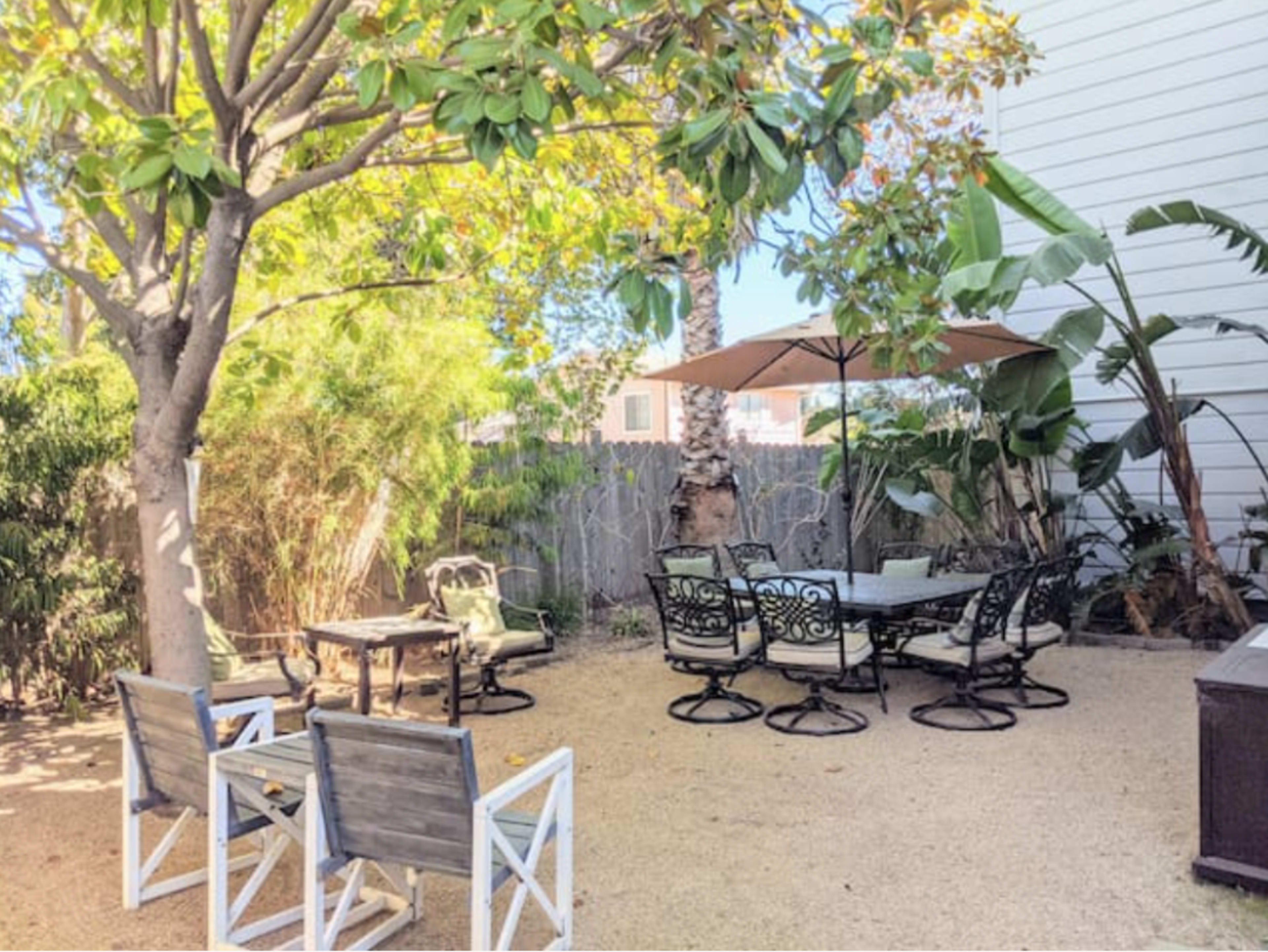 The image shows a backyard patio area with a table and chairs under a large umbrella, surrounded by greenery and a gravel surface.