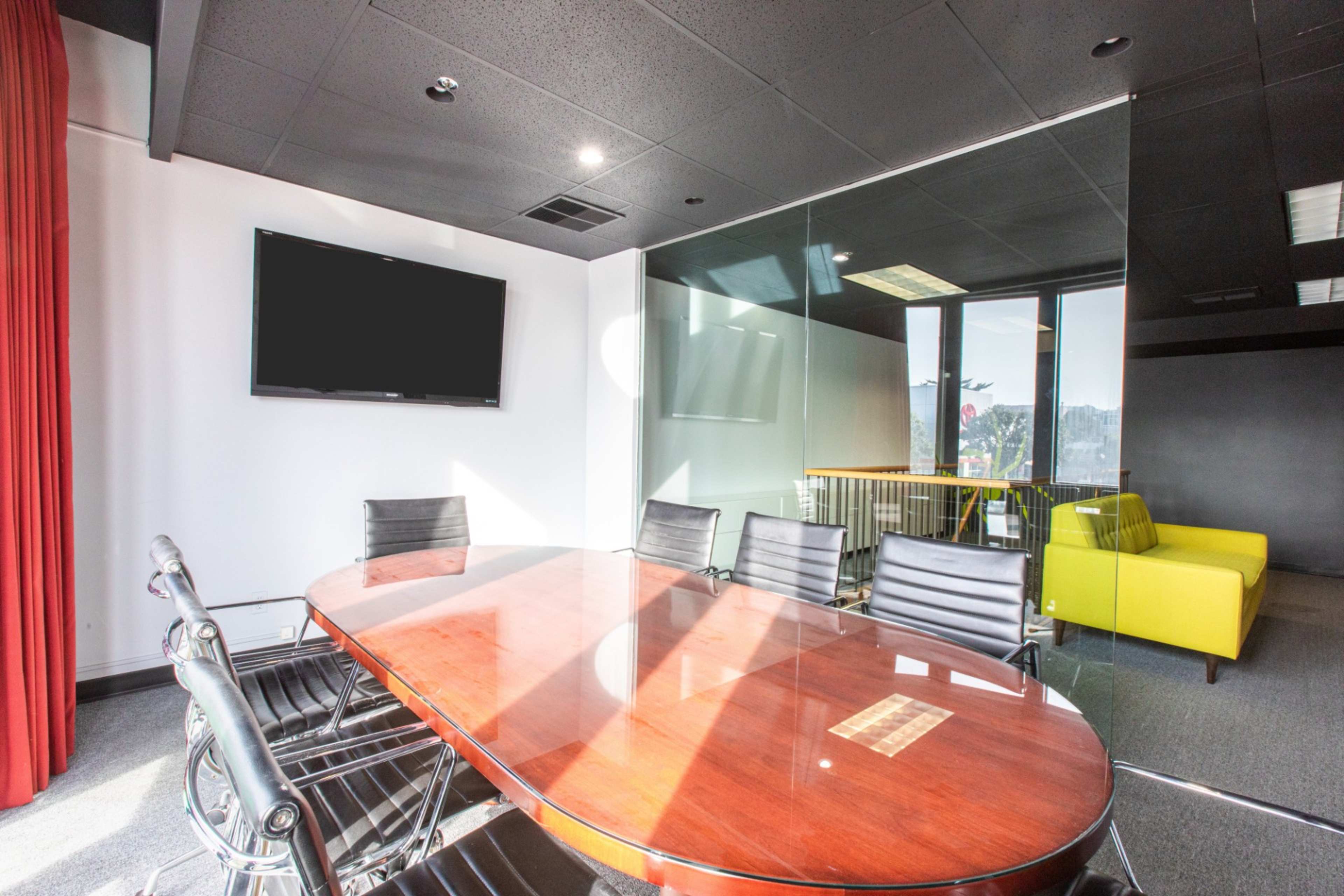 A modern conference room features a wooden oval table surrounded by black chairs, with a large glass wall and a television mounted on the wall.