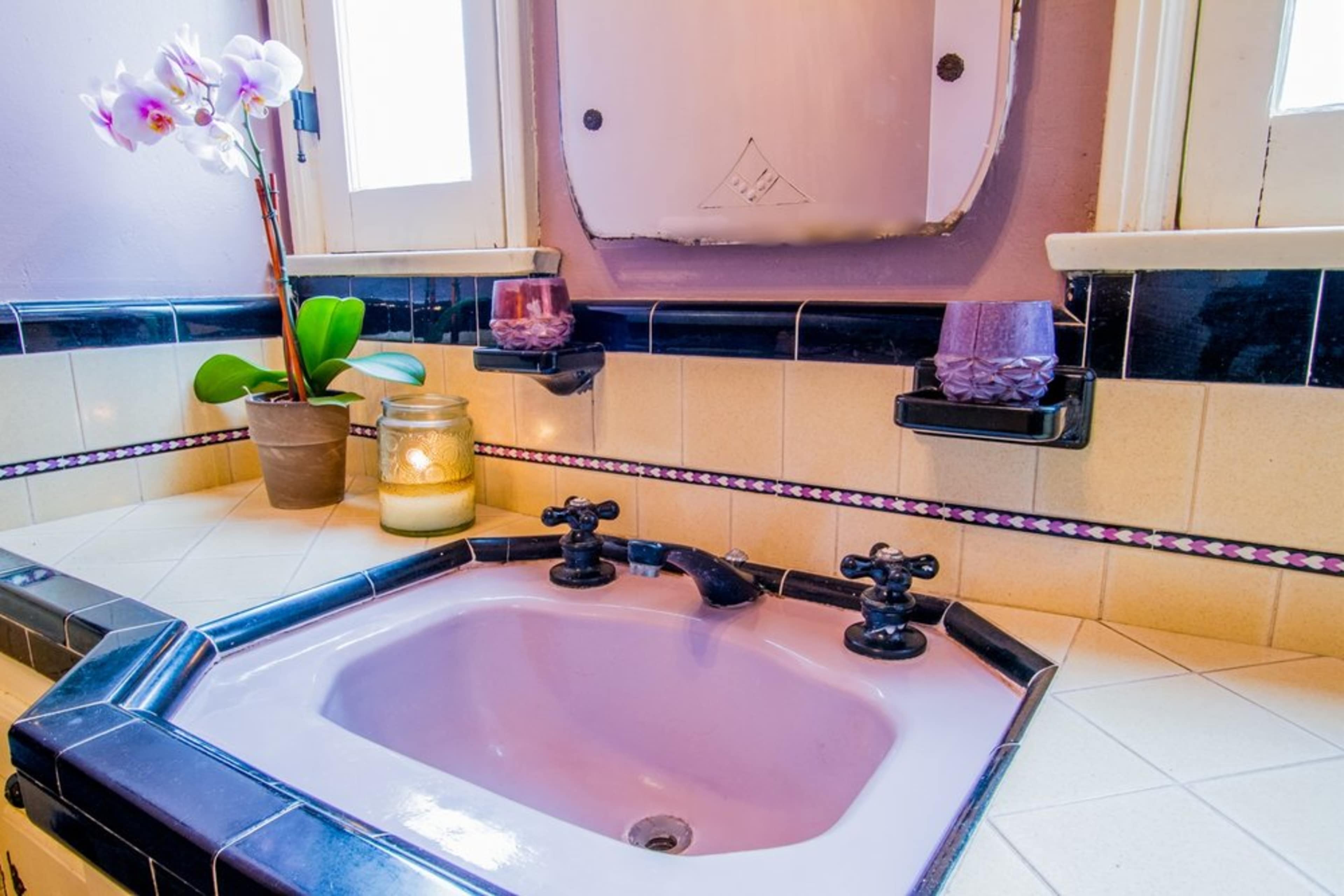 A bathroom countertop with a pink sink, potted orchid, a candle, and decorative soap holders.