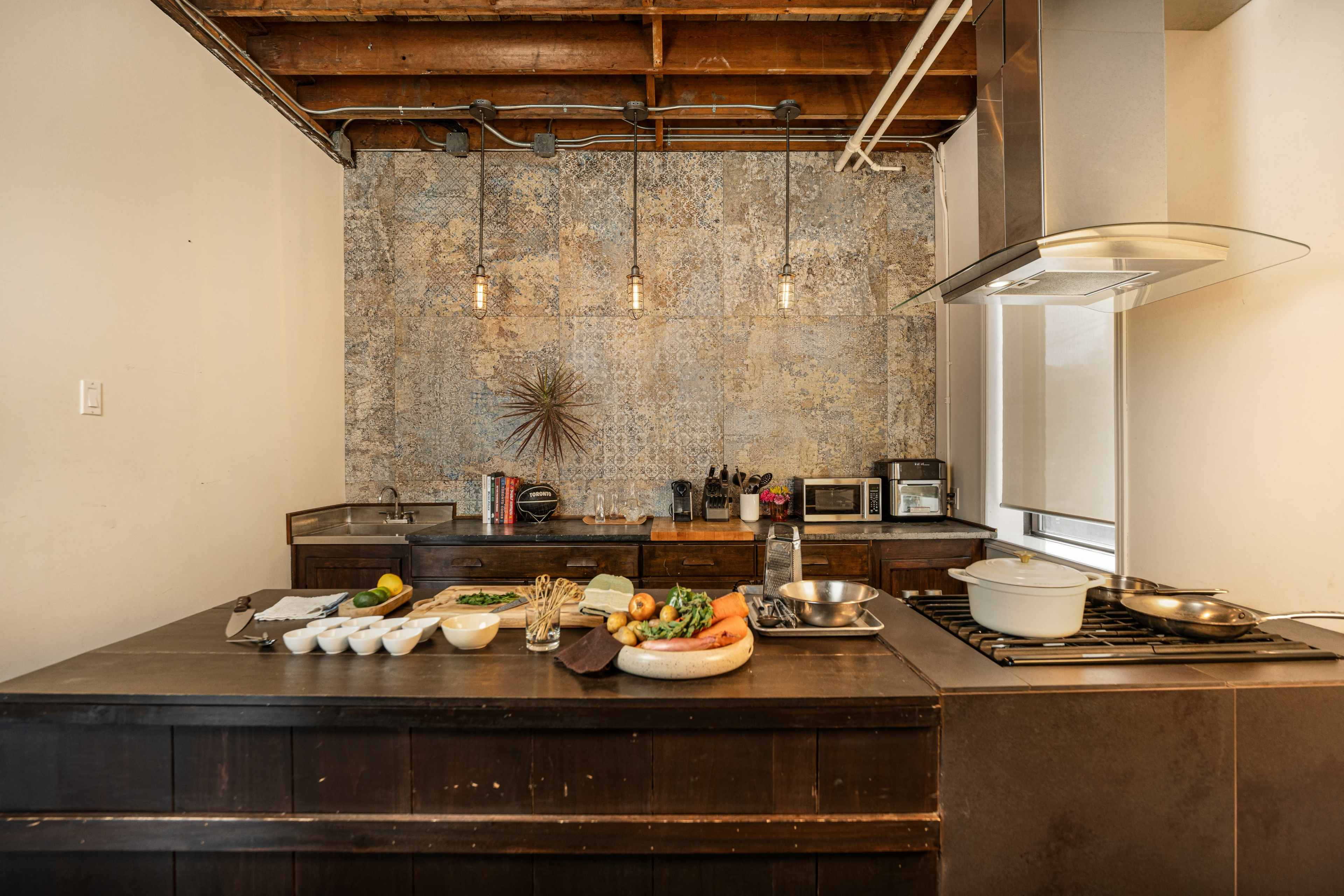 A modern kitchen features a wooden island with various ingredients and cookware arranged on the countertop, illuminated by pendant lights above.
