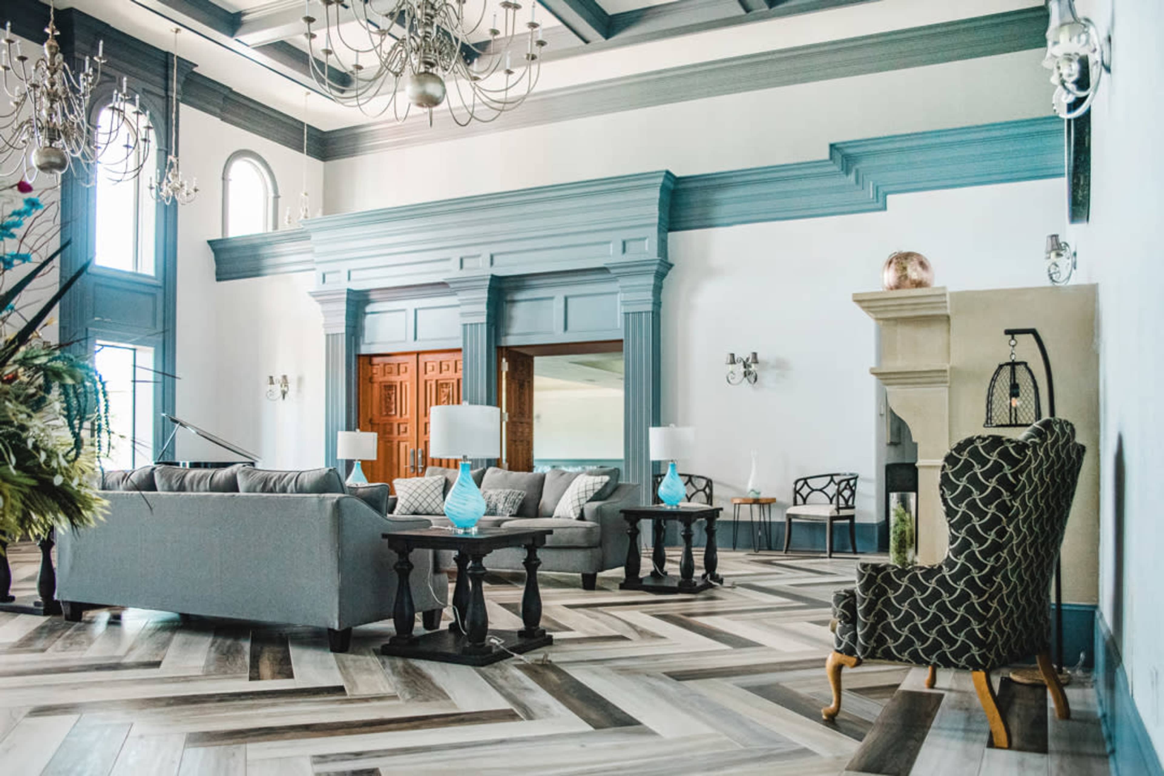 The image shows a spacious, elegantly designed living room with large windows, a prominent fireplace, and a mix of modern and traditional furnishings.