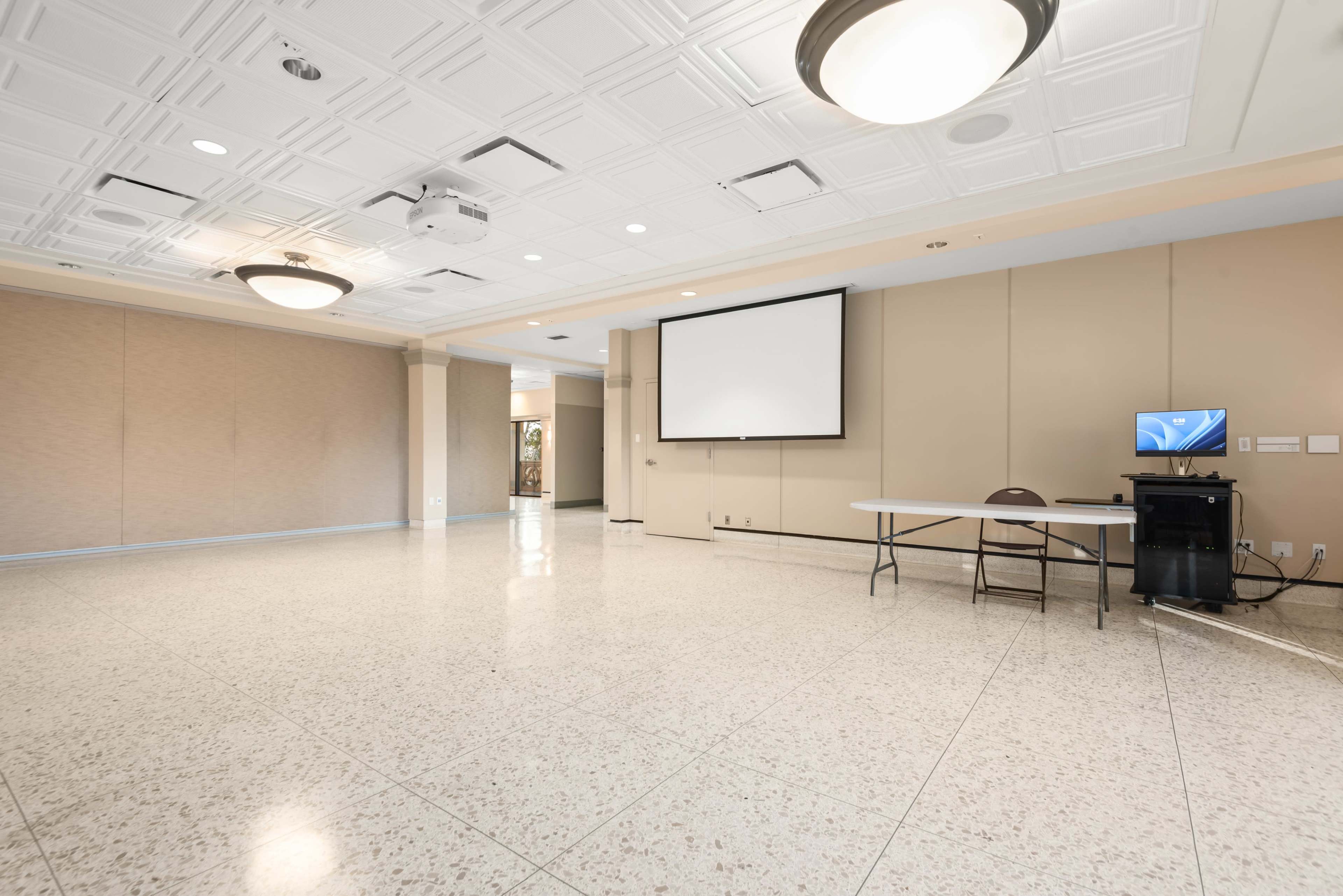The image shows a spacious interior room with a light-colored floor, a projector screen mounted on the wall, a table, and a computer setup on a cart.