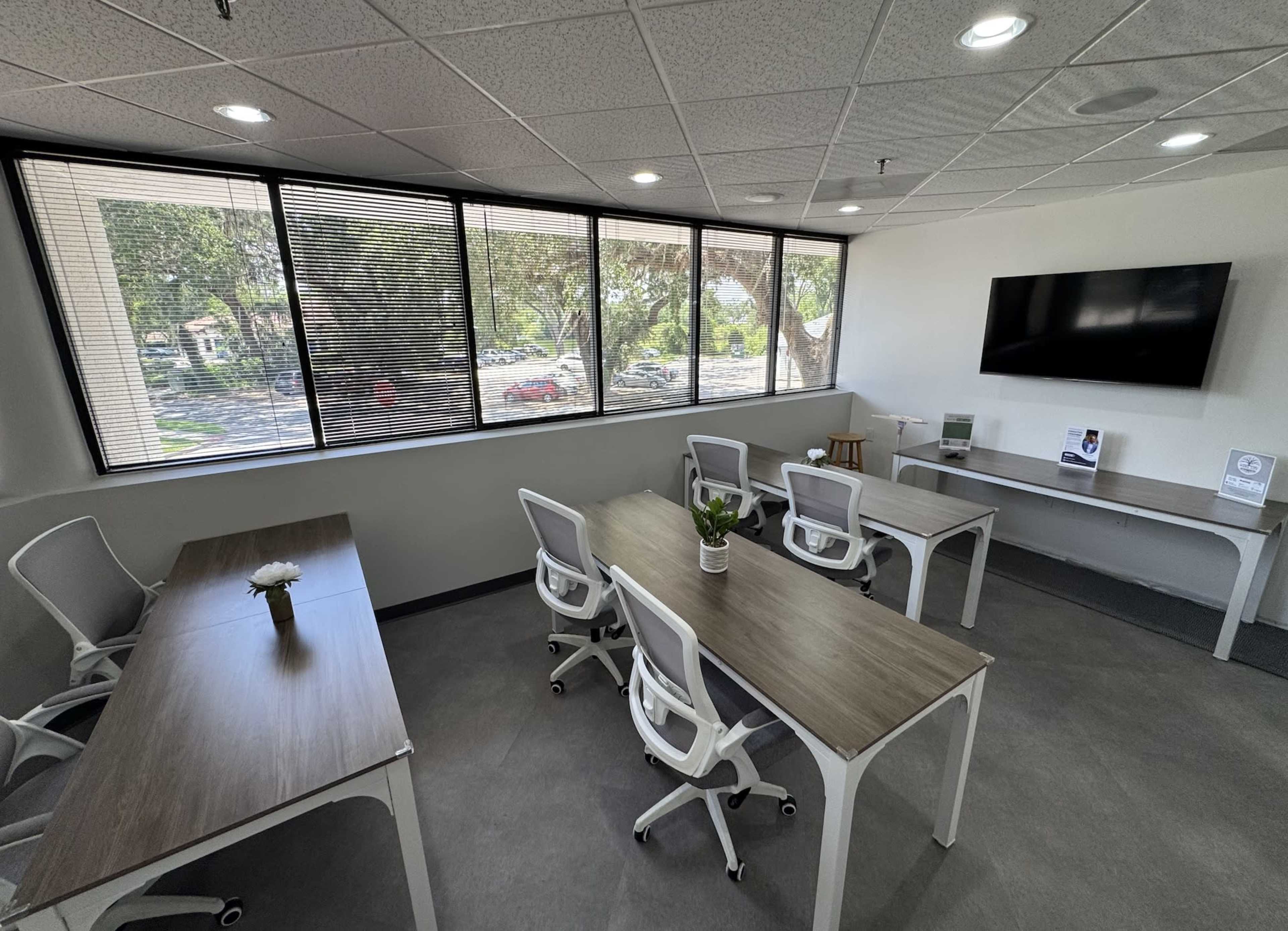 The image shows a modern office space featuring several workstations with desks, chairs, and a large window, complemented by a television on the wall.