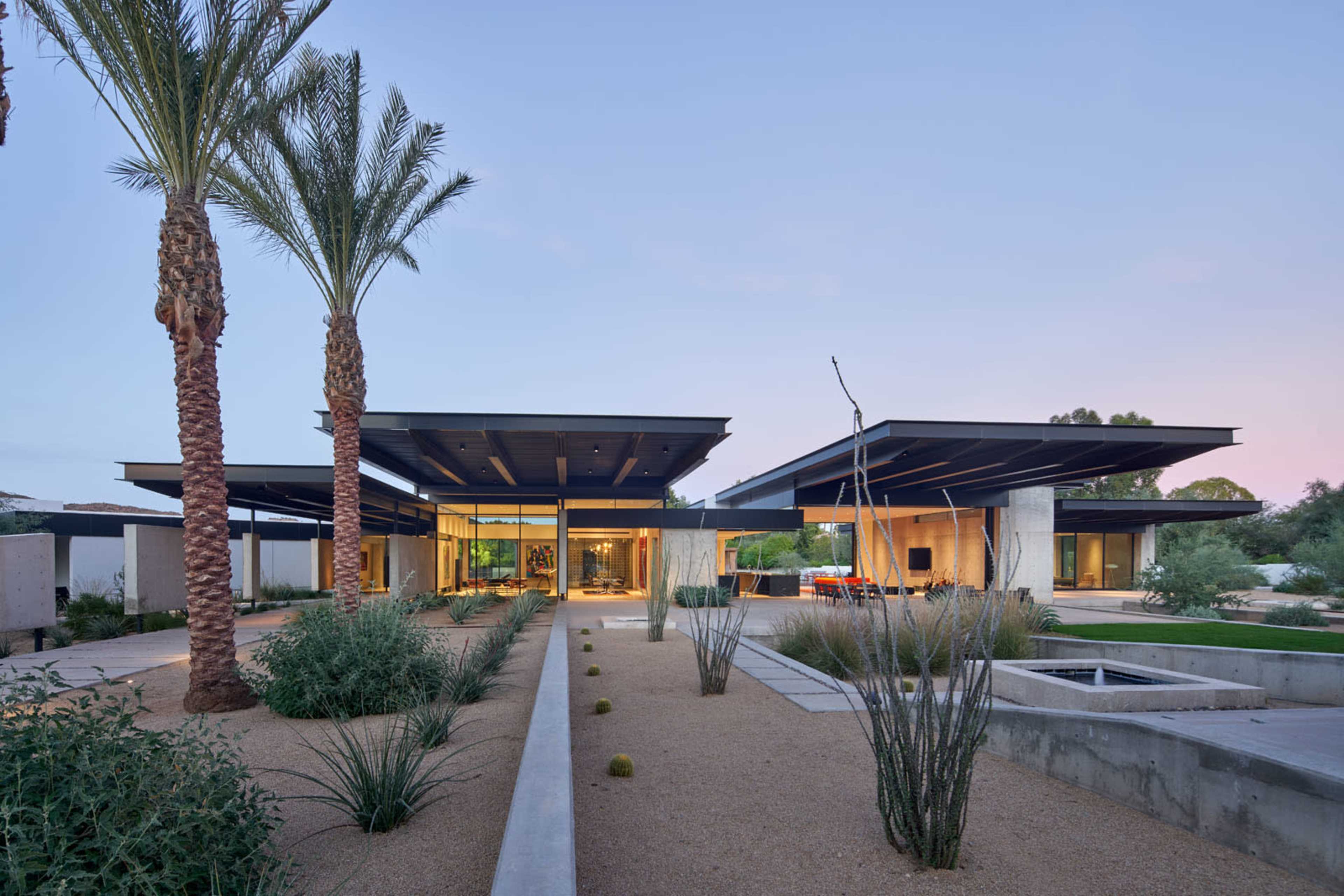 A modern, single-story house features expansive flat roofs and is surrounded by desert landscaping, including palm trees and low shrubs.