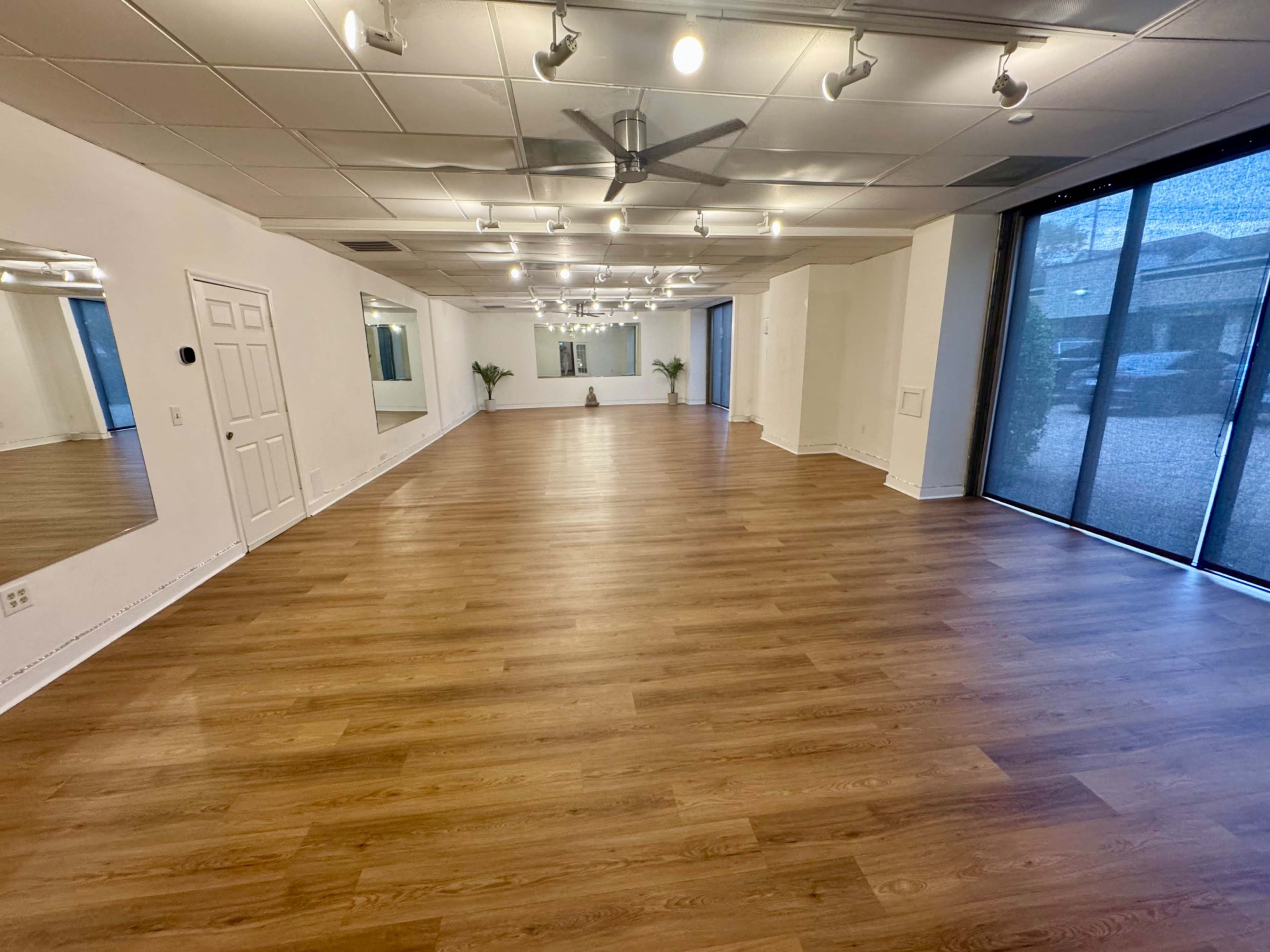 The image shows a spacious, empty interior room with wooden flooring, bright lighting from overhead fixtures, and large windows letting in natural light.