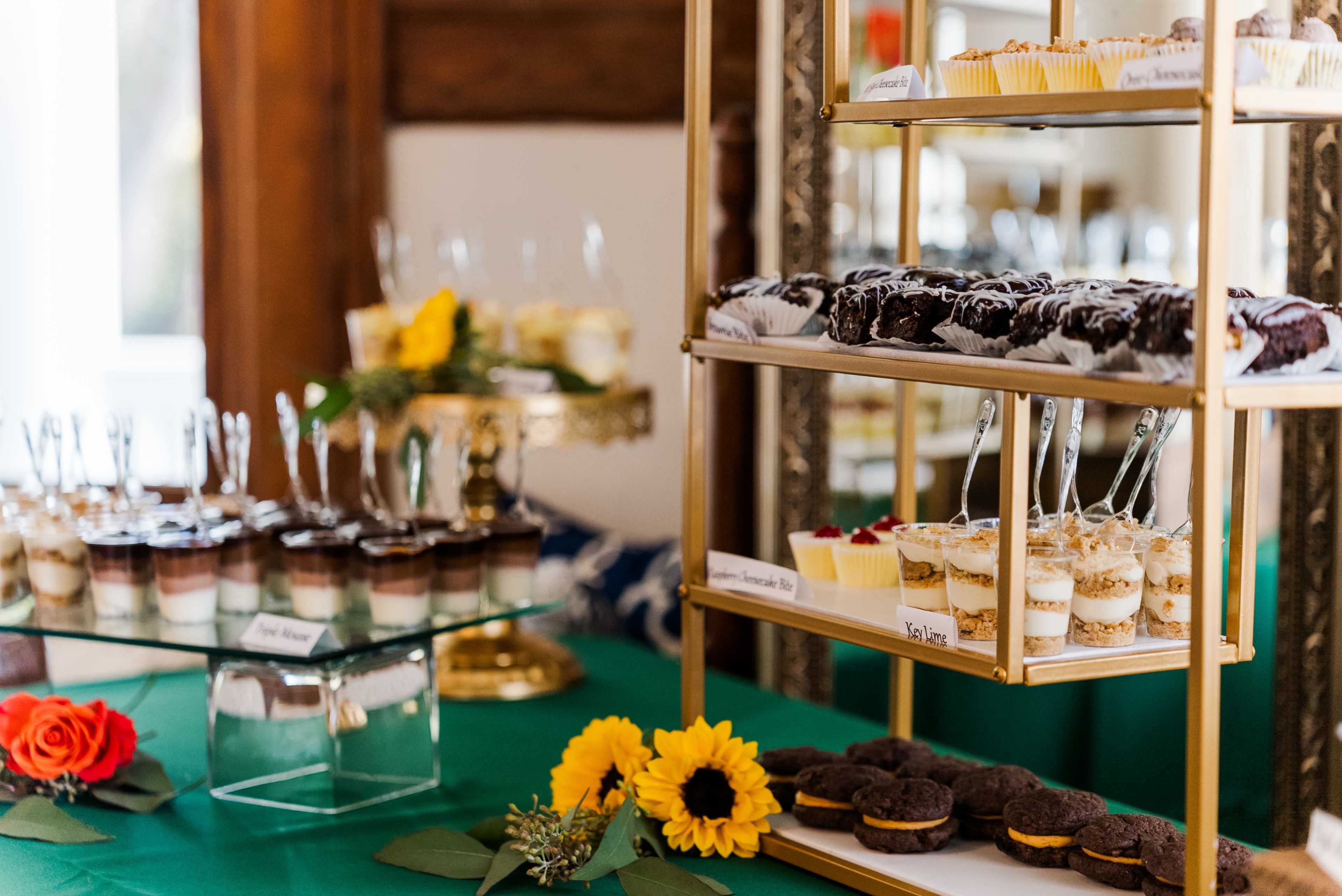The display features a three-tiered stand filled with various desserts, including cupcakes, layered parfaits, and cookies, arranged alongside fresh flowers and greenery.
