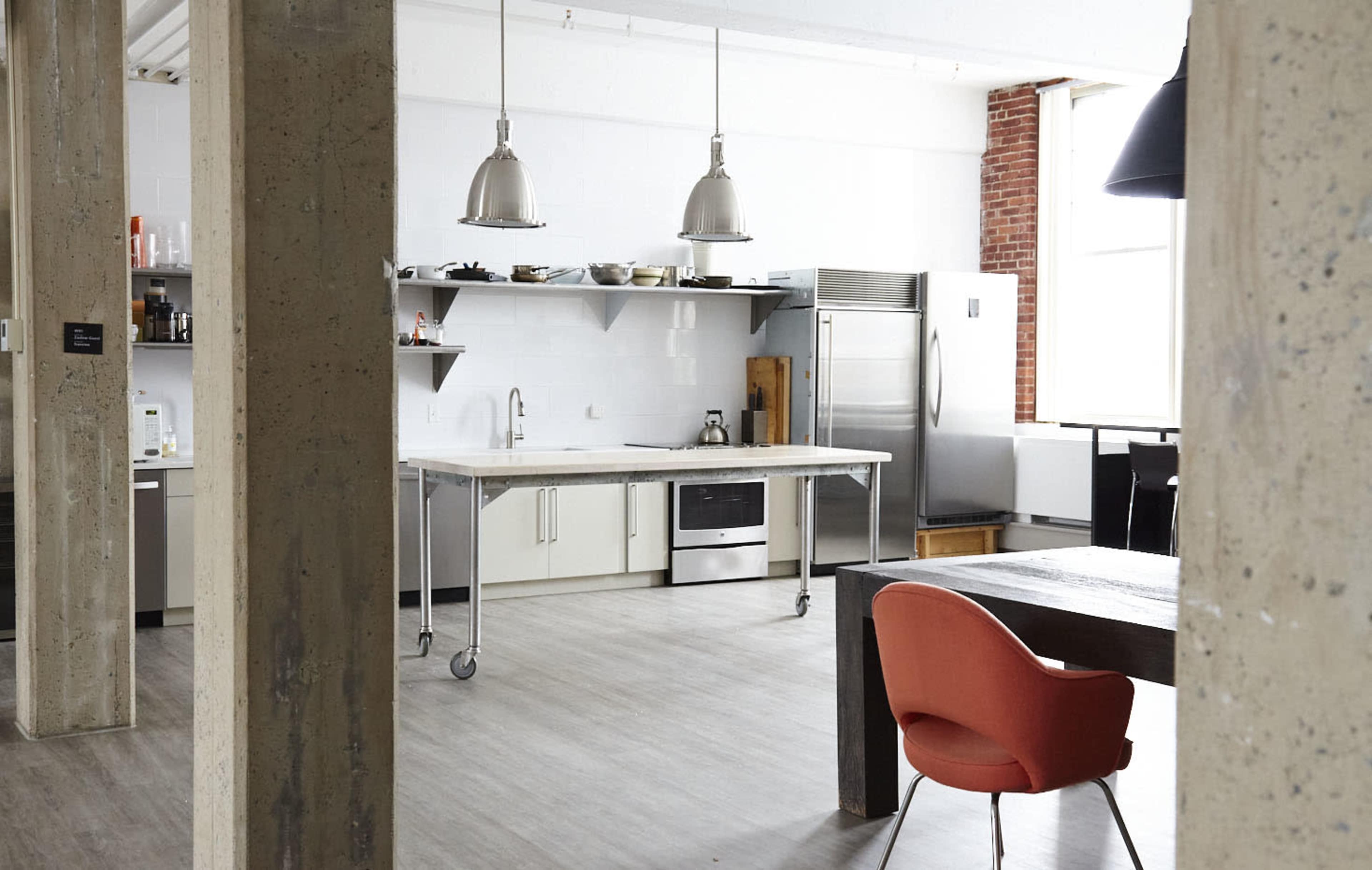 The image shows a modern kitchen with stainless steel appliances, a central island, and a dining area featuring a wooden table and an orange chair.