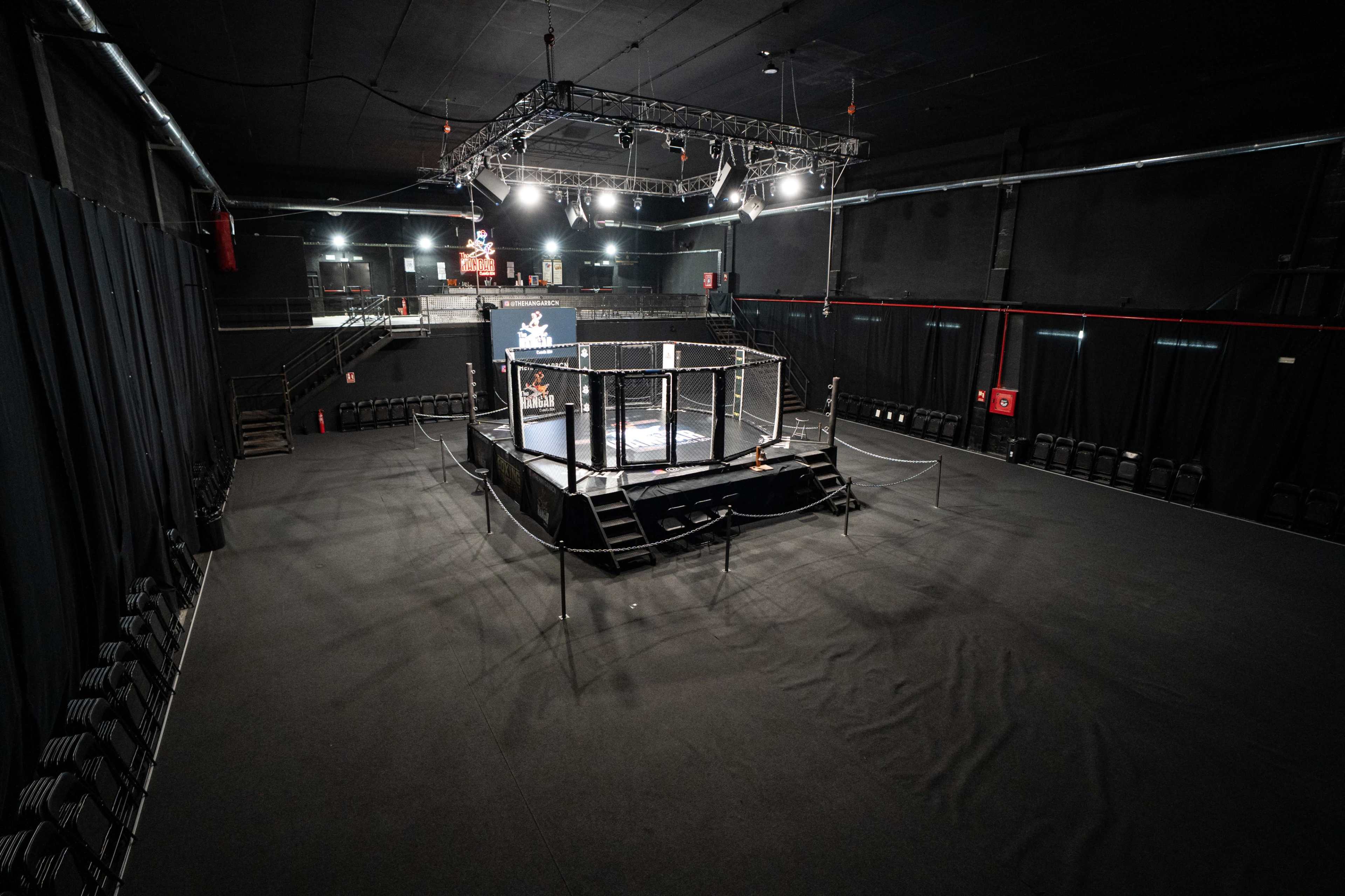 The image shows an empty indoor arena featuring a central cage-like structure, surrounded by dark curtains and rows of unoccupied seats.