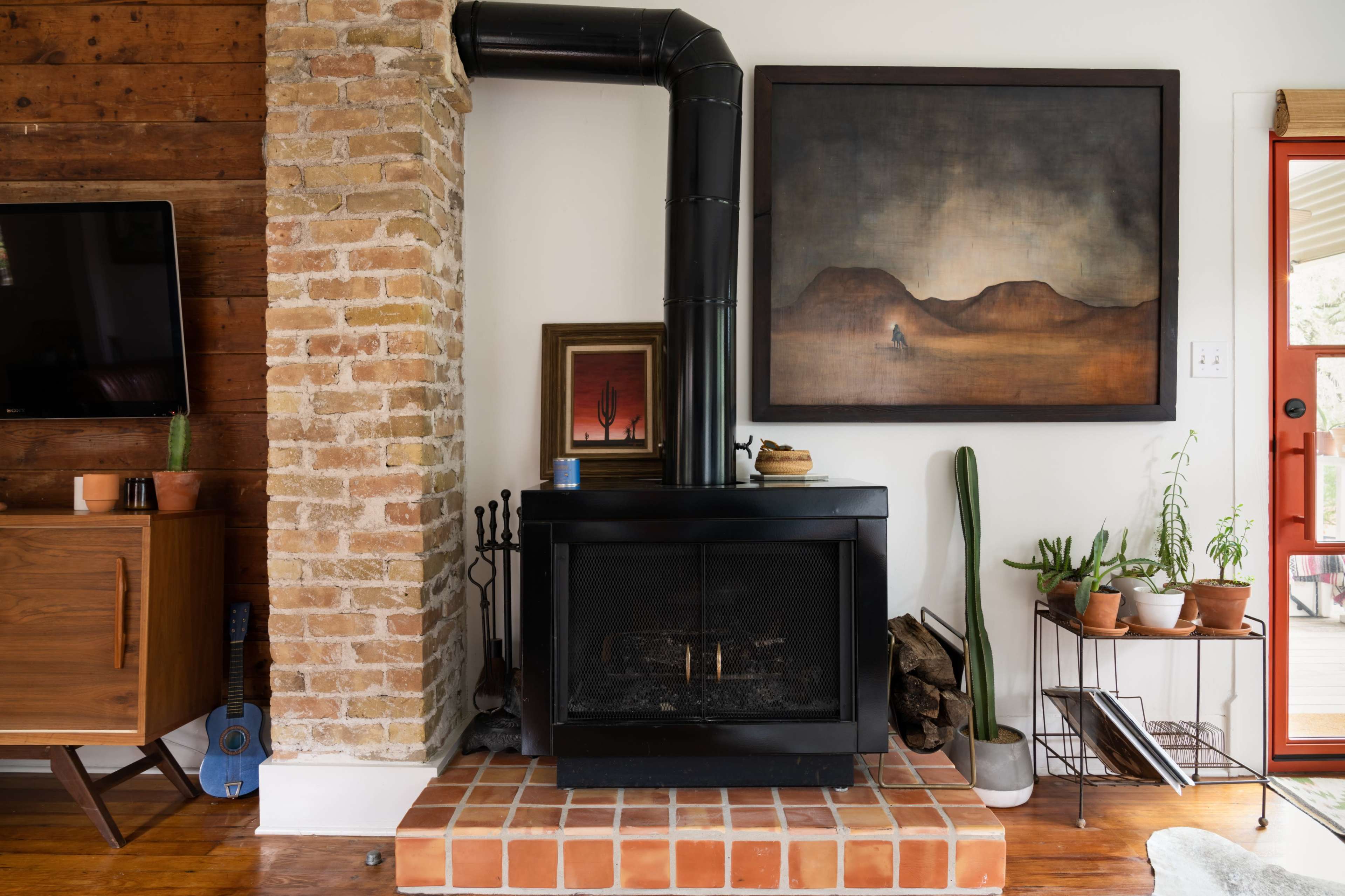 A living room with a brick fireplace and a black wood stove, accompanied by a large landscape painting and plant arrangements.