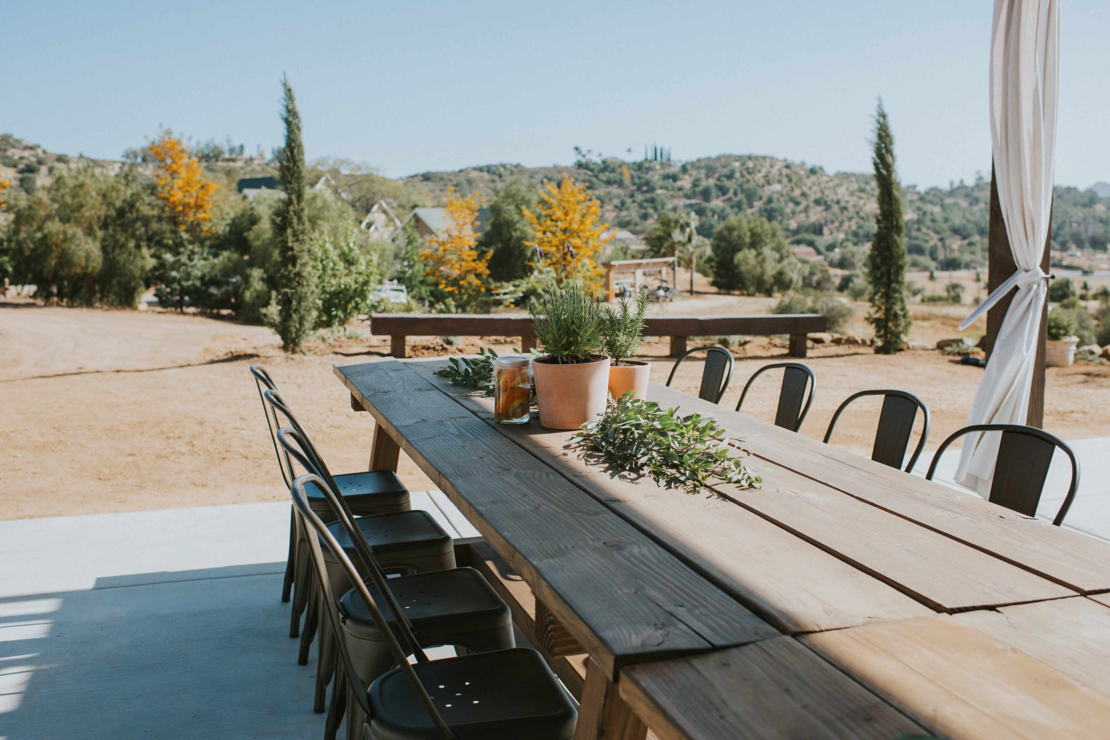 A long wooden table with black chairs is set outside, surrounded by greenery and a rustic landscape in the background.
