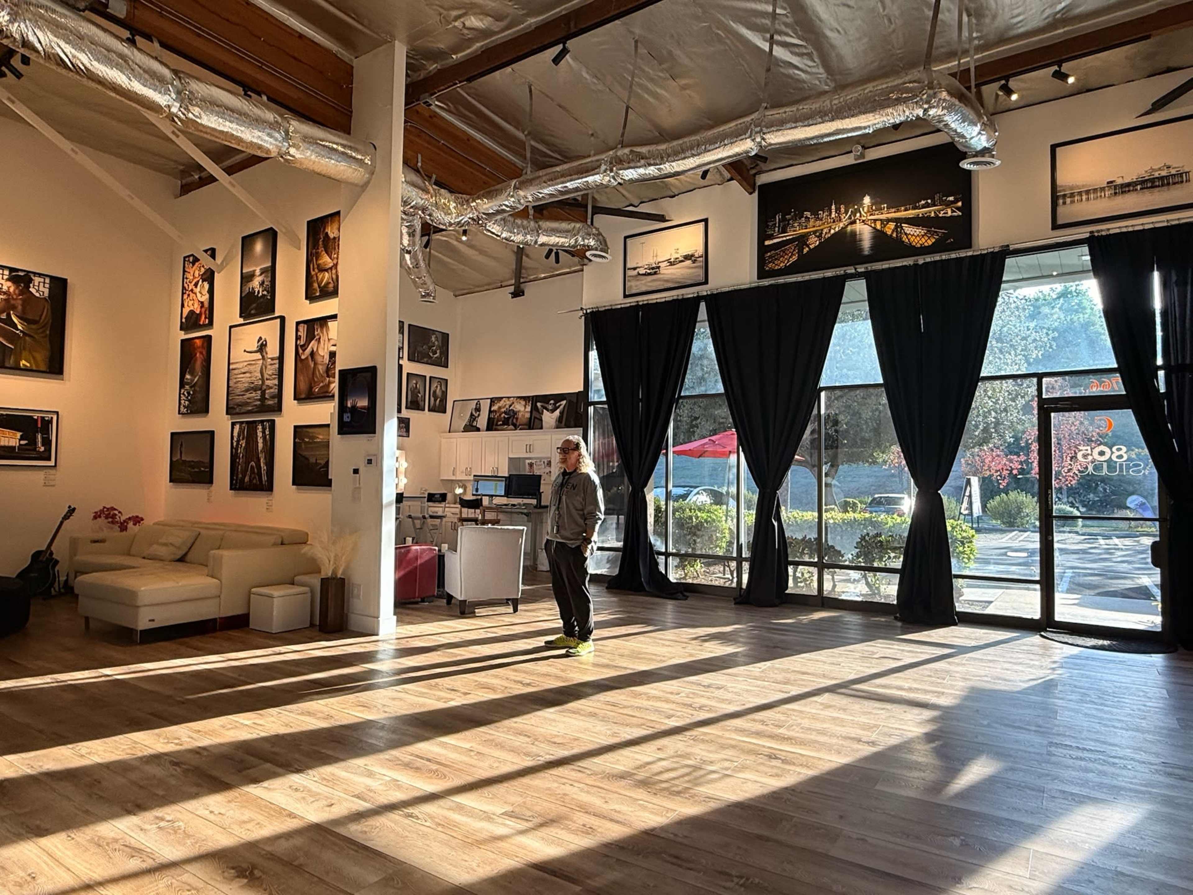 An empty art gallery features large windows, a white sofa, and black drapes, with a lone figure standing in the sunlight casting long shadows on the wooden floor.