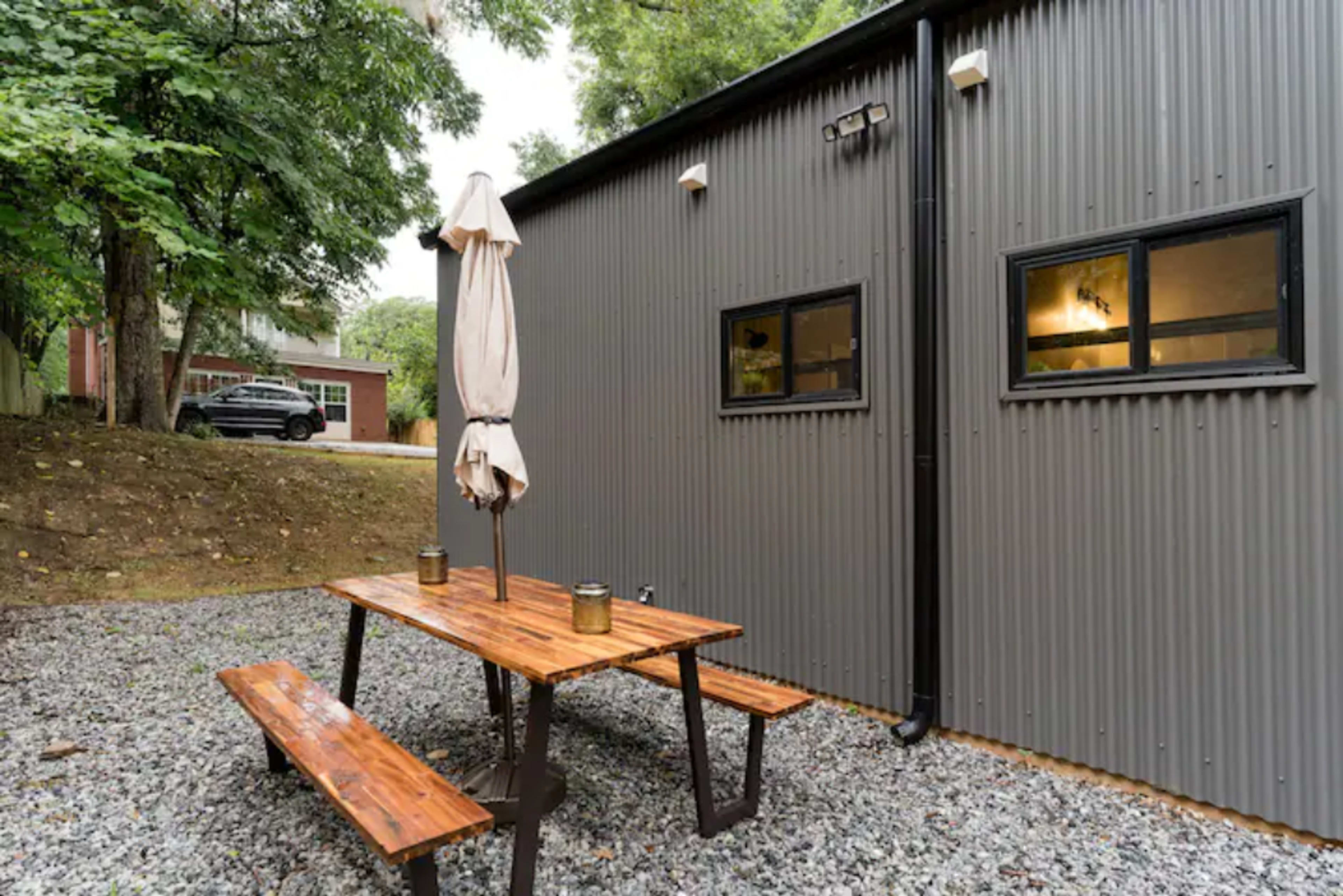 A wooden picnic table with benches and a shade umbrella is positioned on gravel in front of a gray, corrugated metal building surrounded by trees.