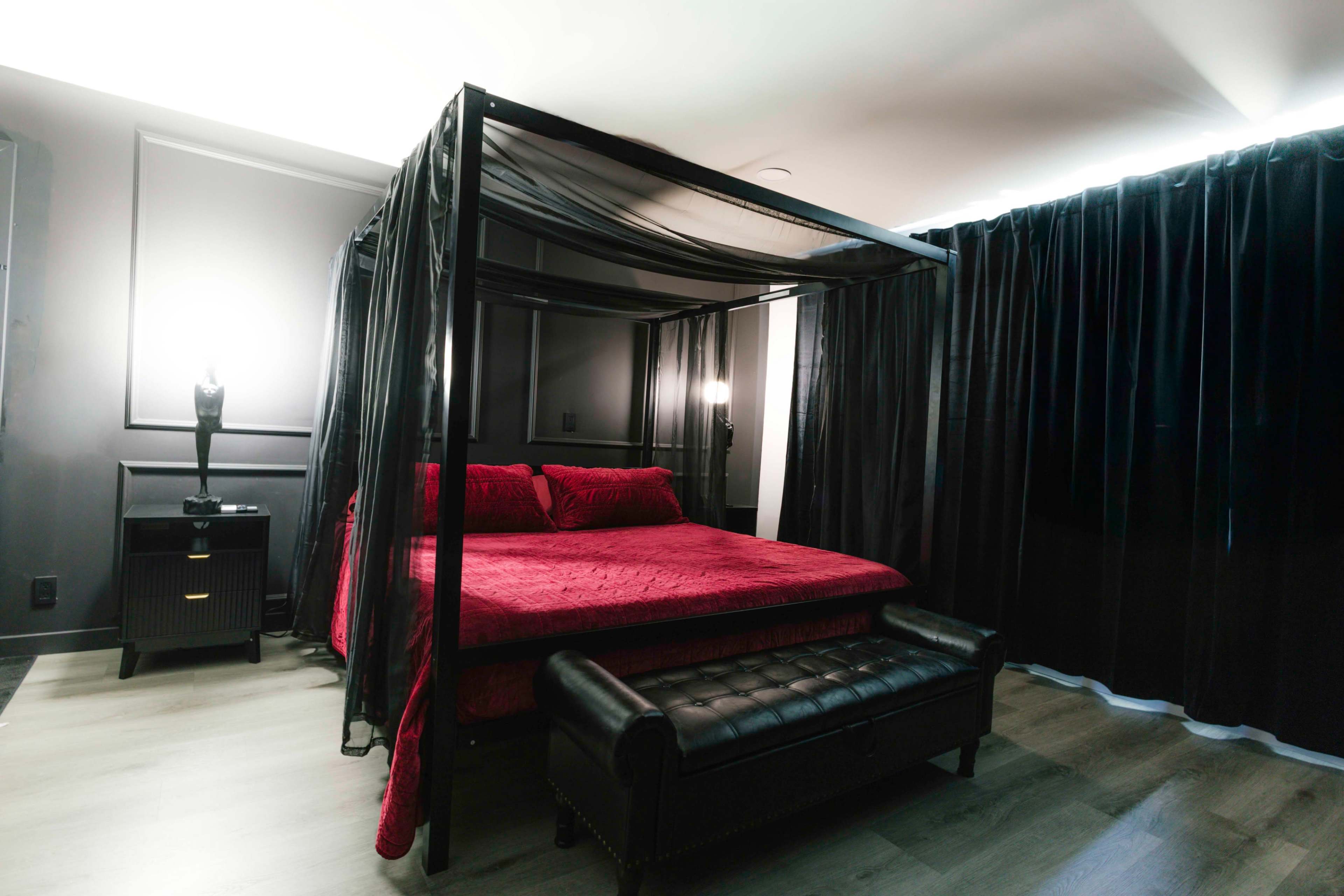 The image shows a bedroom featuring a black four-poster bed with a red quilt and a black bench at the foot, surrounded by dark curtains and minimal lighting.