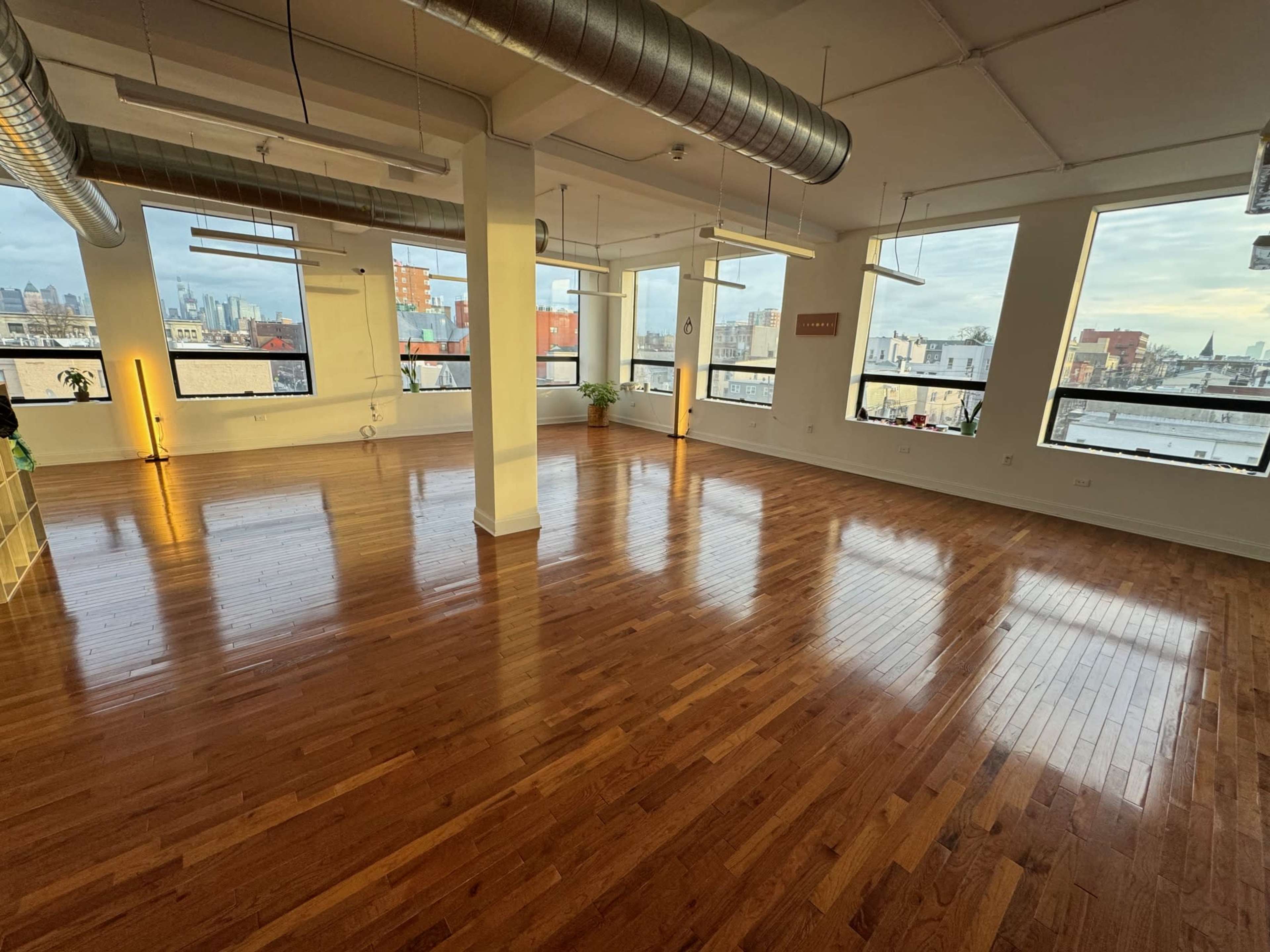 The image shows a spacious, empty room with polished wooden floors and large windows offering views of the city skyline.