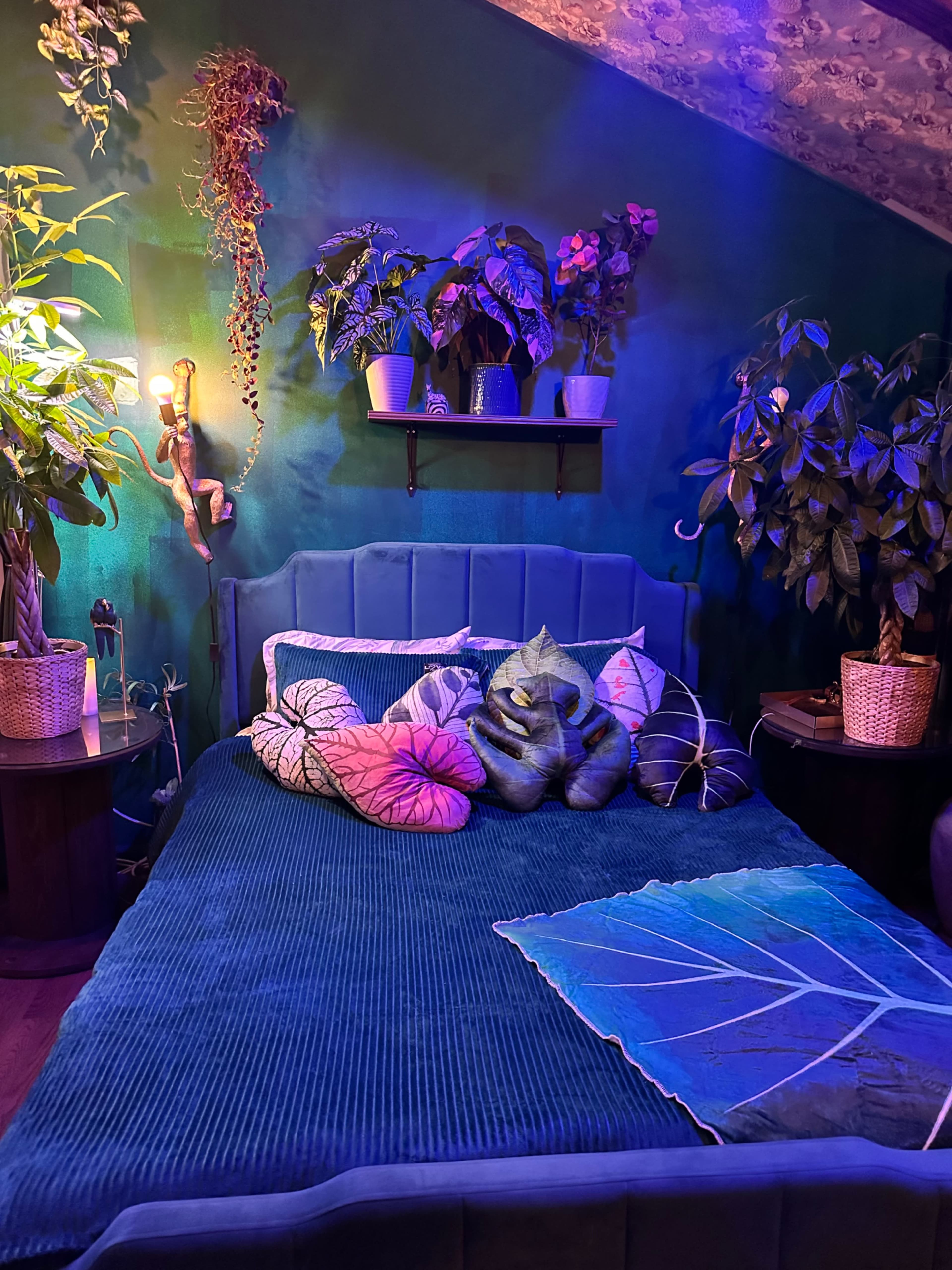 A cozy bedroom features a green wall, a plush bed adorned with colorful leaf-patterned pillows, and various plants arranged on shelves and around the room.