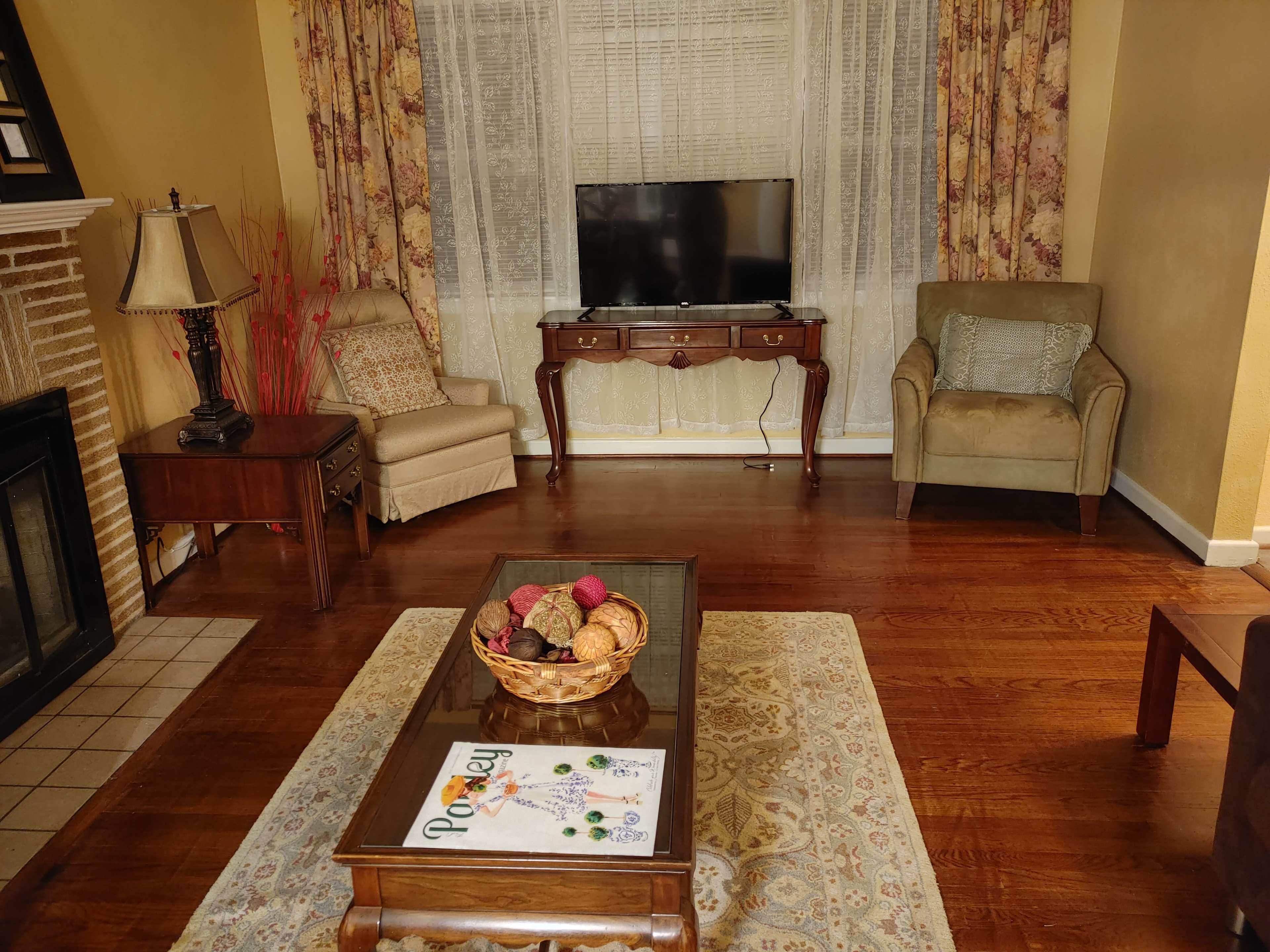 The living room features two chairs, a wooden coffee table with a decorative bowl, a TV stand with a flat-screen television, and a light-colored rug on polished wooden floors.