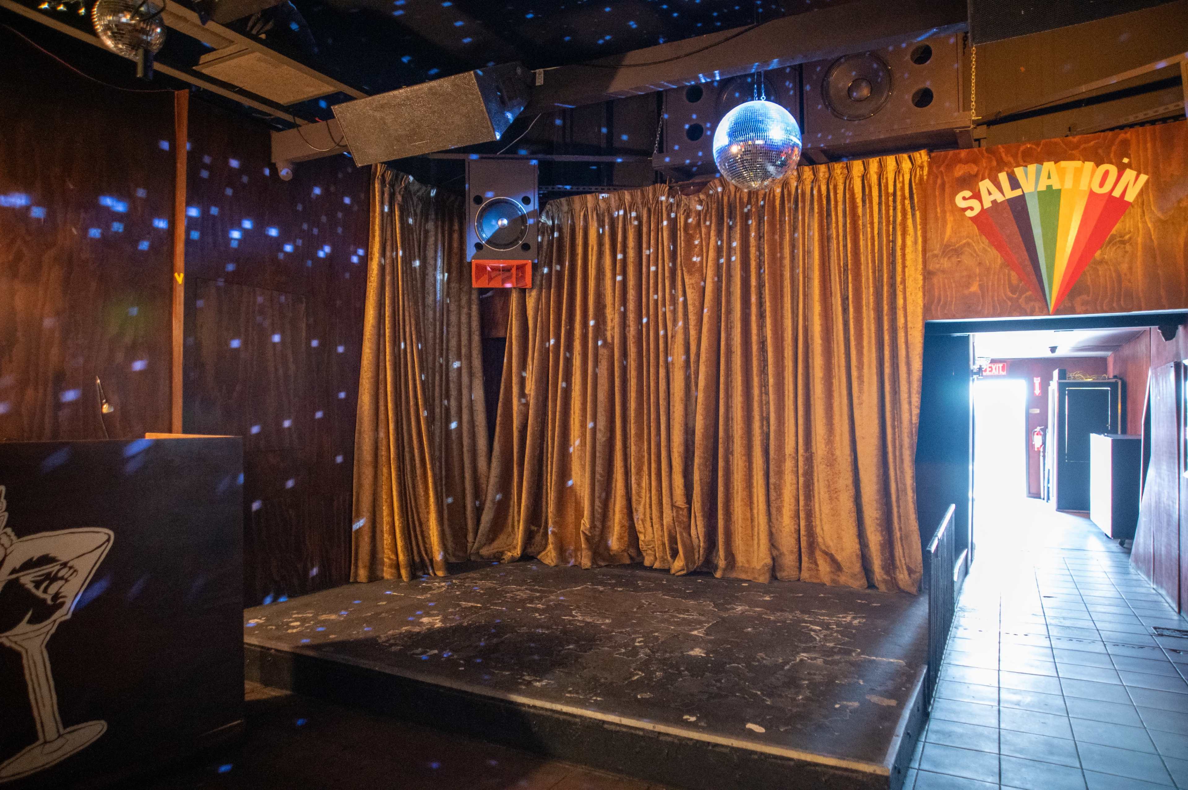 The image shows a dimly lit performance stage with golden curtains, a disco ball, and a doorway leading out of the venue.