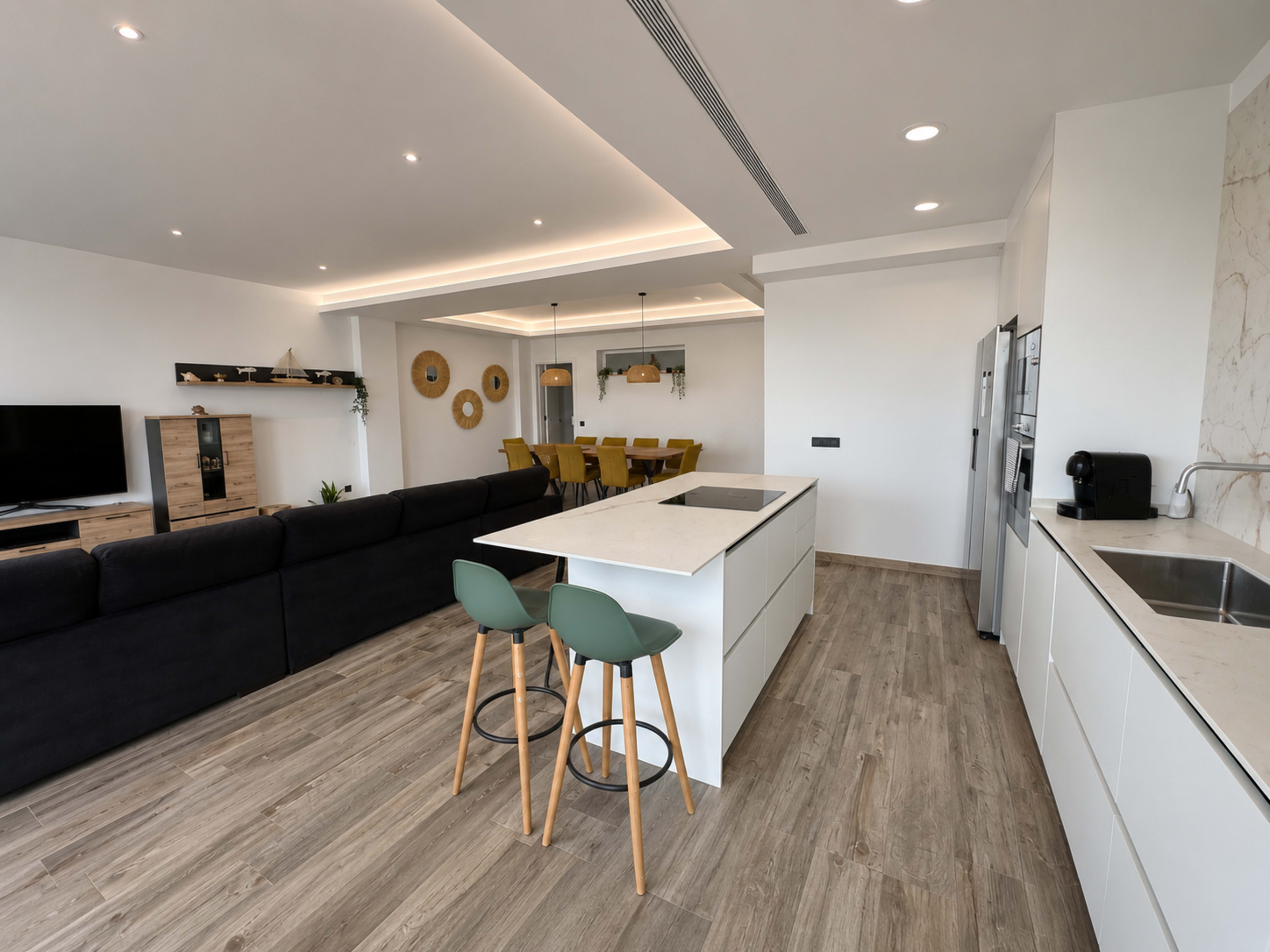 The image shows a modern open-concept kitchen and living area with a central island, seating, and a dining table in the background.