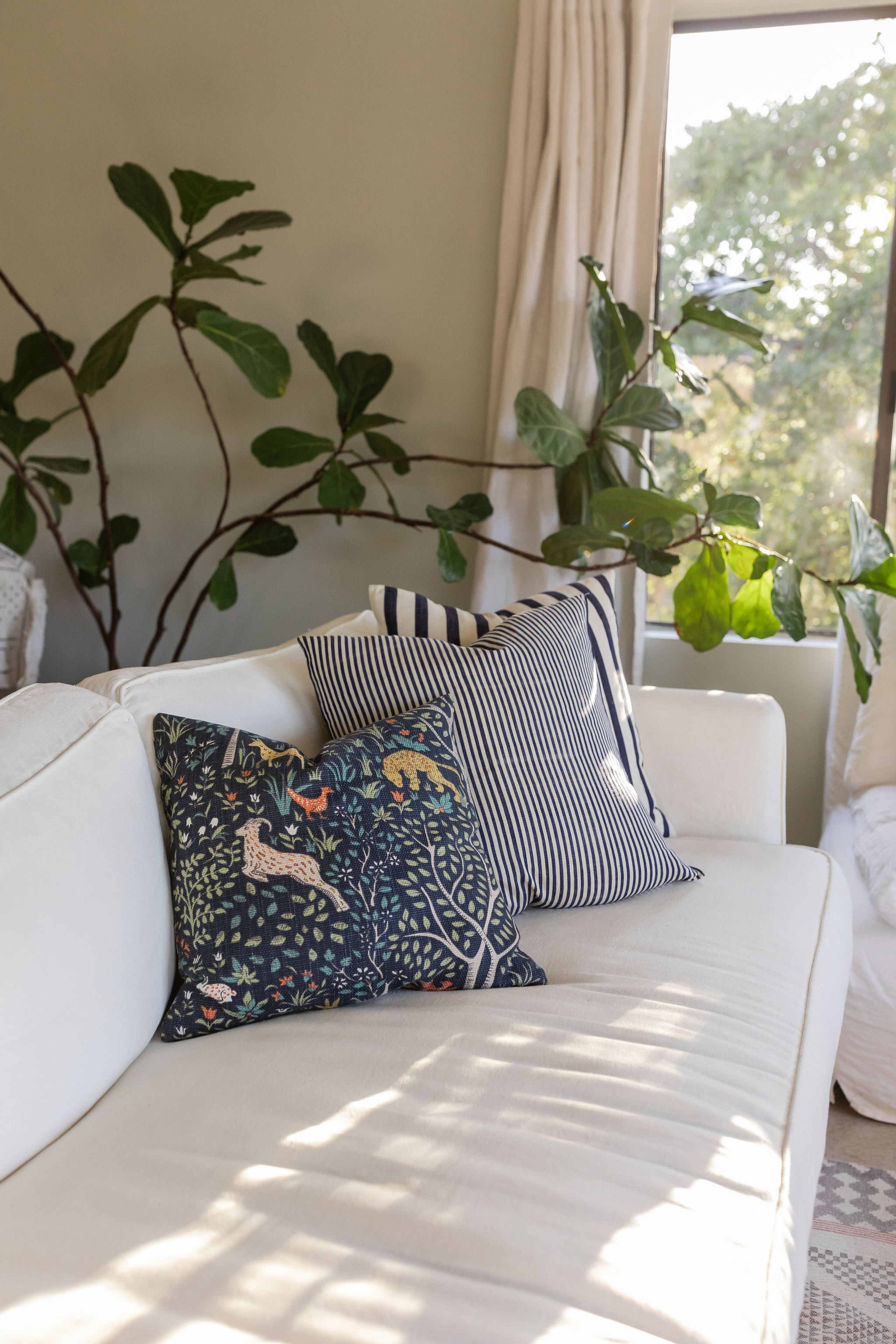 A white sofa with decorative pillows is positioned beside a large window with greenery visible outside.