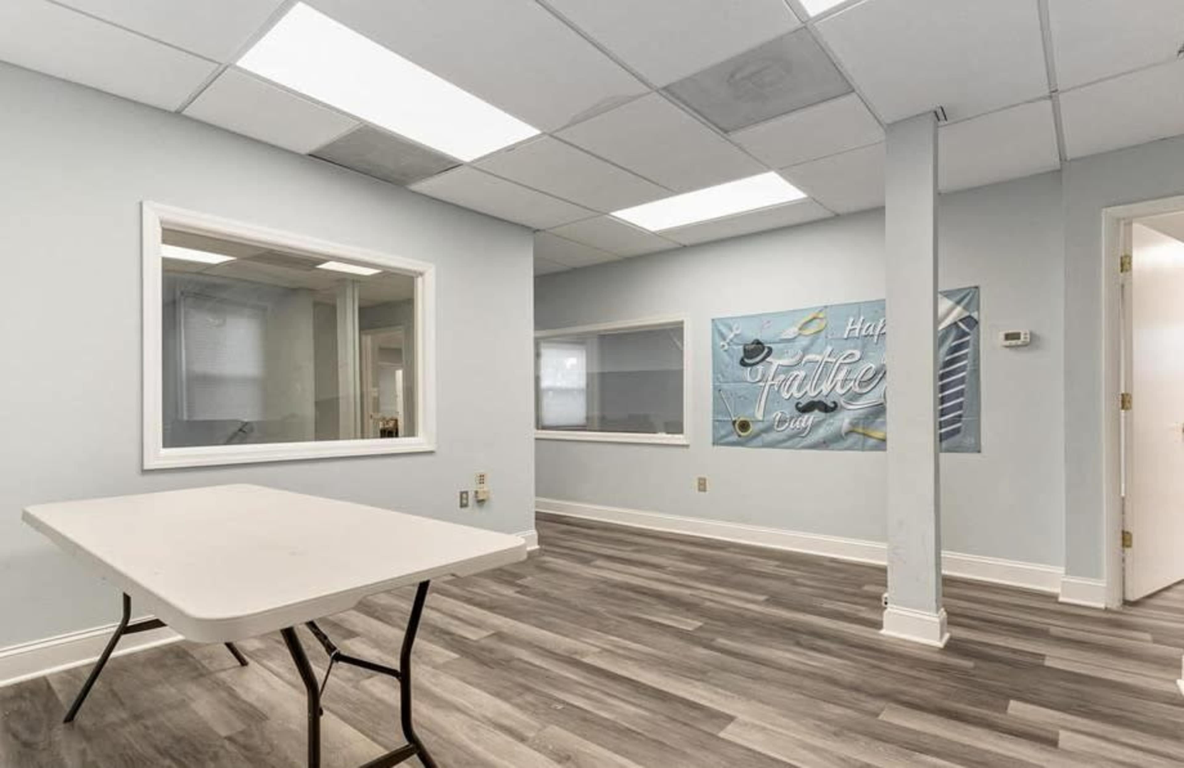 A sparsely furnished office space features a table, gray walls, and large windows with a wall mural that reads "Half Baked Cafe."