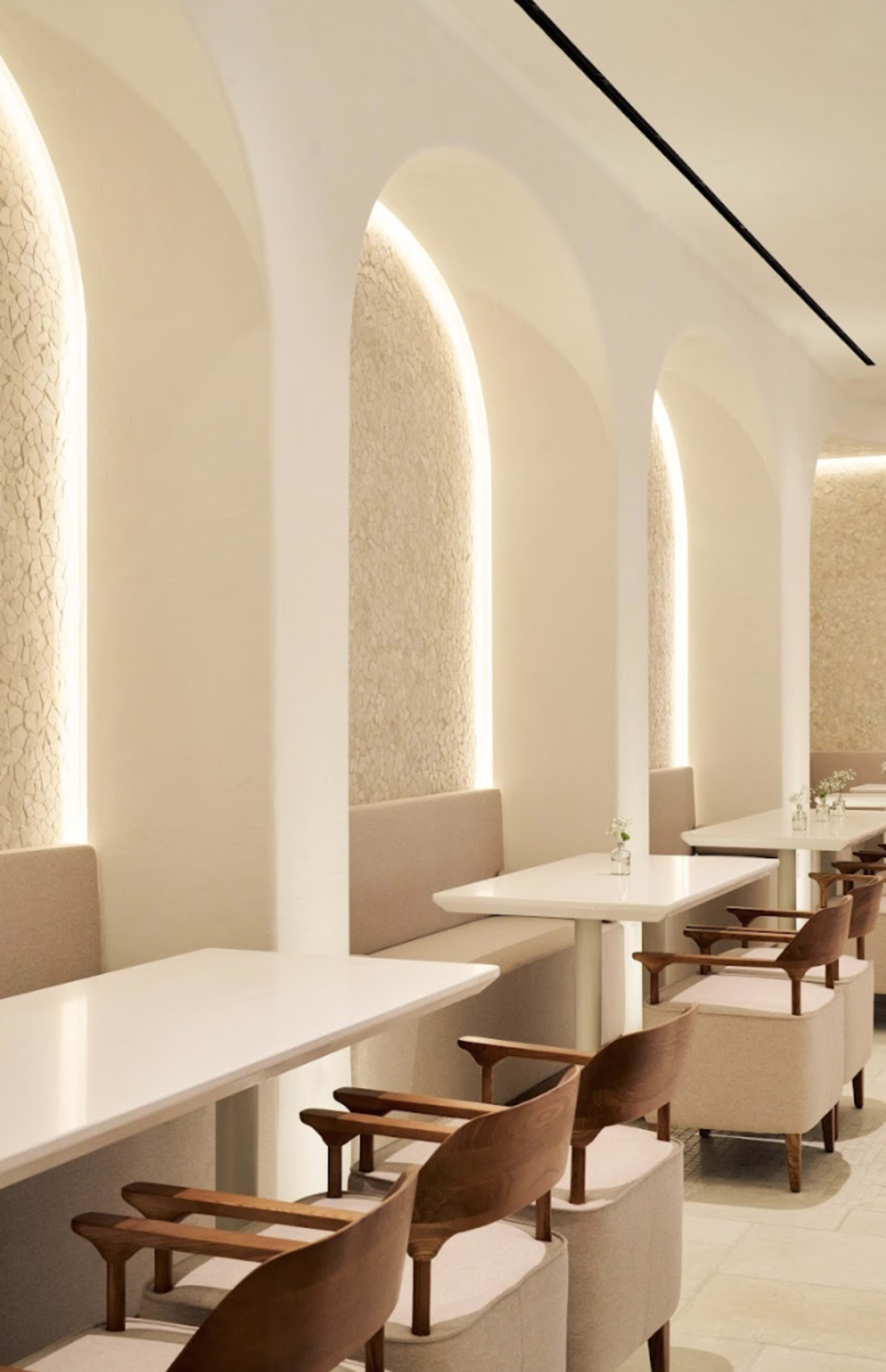 The image shows a modern café interior with white arches, minimalistic wooden chairs, and tables evenly spaced along the walls.